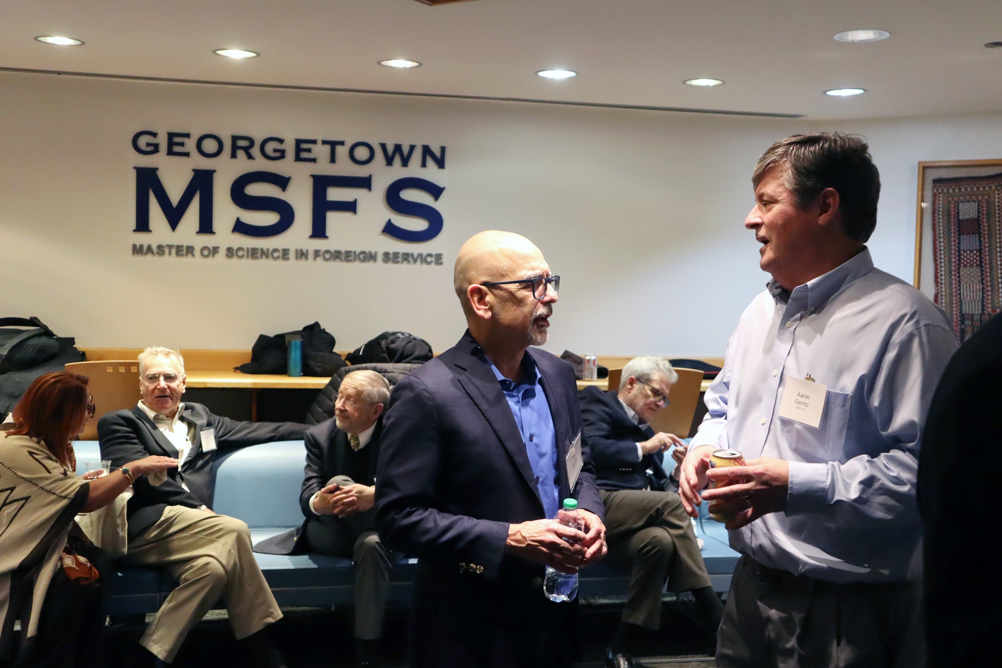 People standing and sitting in a lounge, networking. The wall has a sign that says "Georgetown MSFS, Master of Science in Foreign Service"