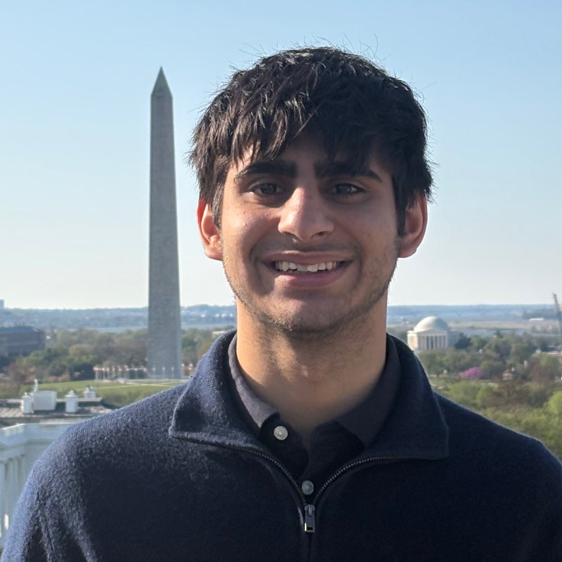 Headshot of Riz KHAN smiling, behind him the Washington monument