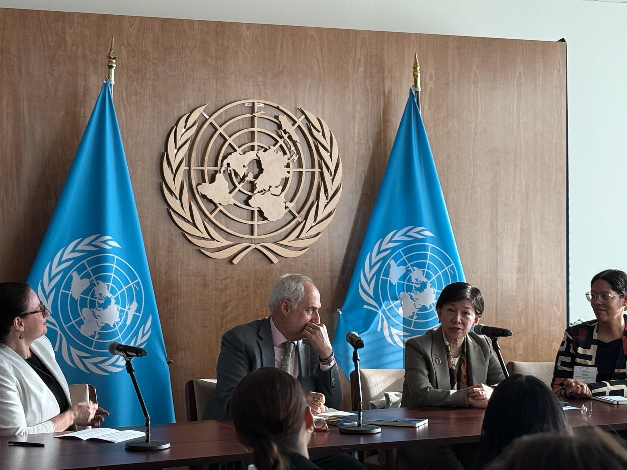 Four people engaging in a panel discussion at the United Nations Headquarters. Behind them the logo for the UN.