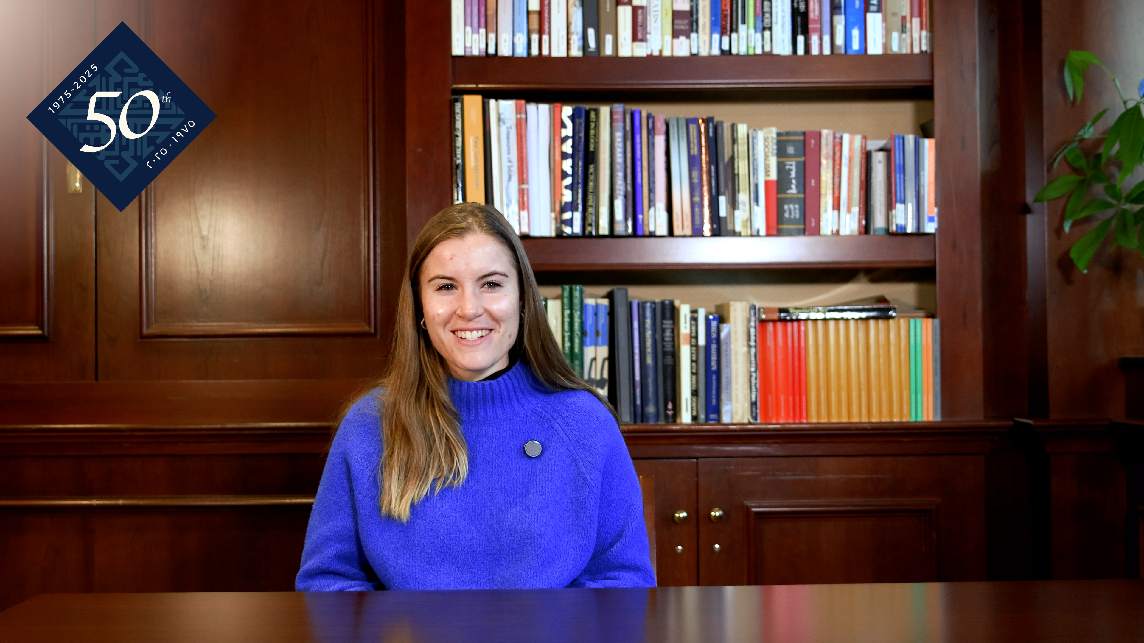 A person wearing a blue sweater sits smiling in front of a bookshelf filled with colorful books. There is a "50th Anniversary" logo in the upper left corner.