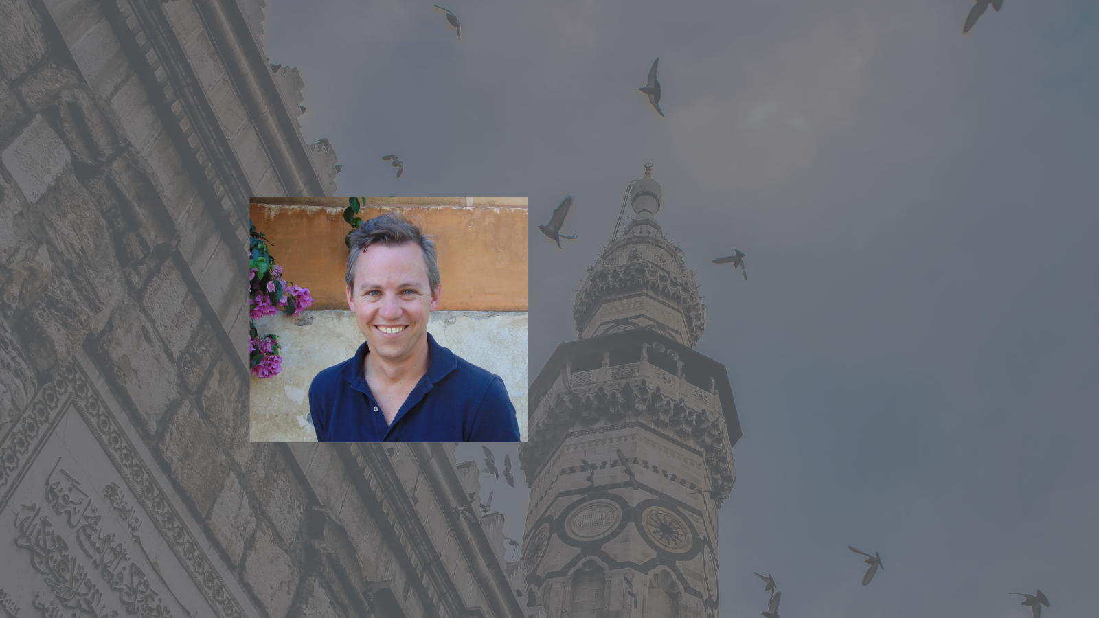A man in a dark blue polo smiles agains a stucco background with small purple flowers to his right.