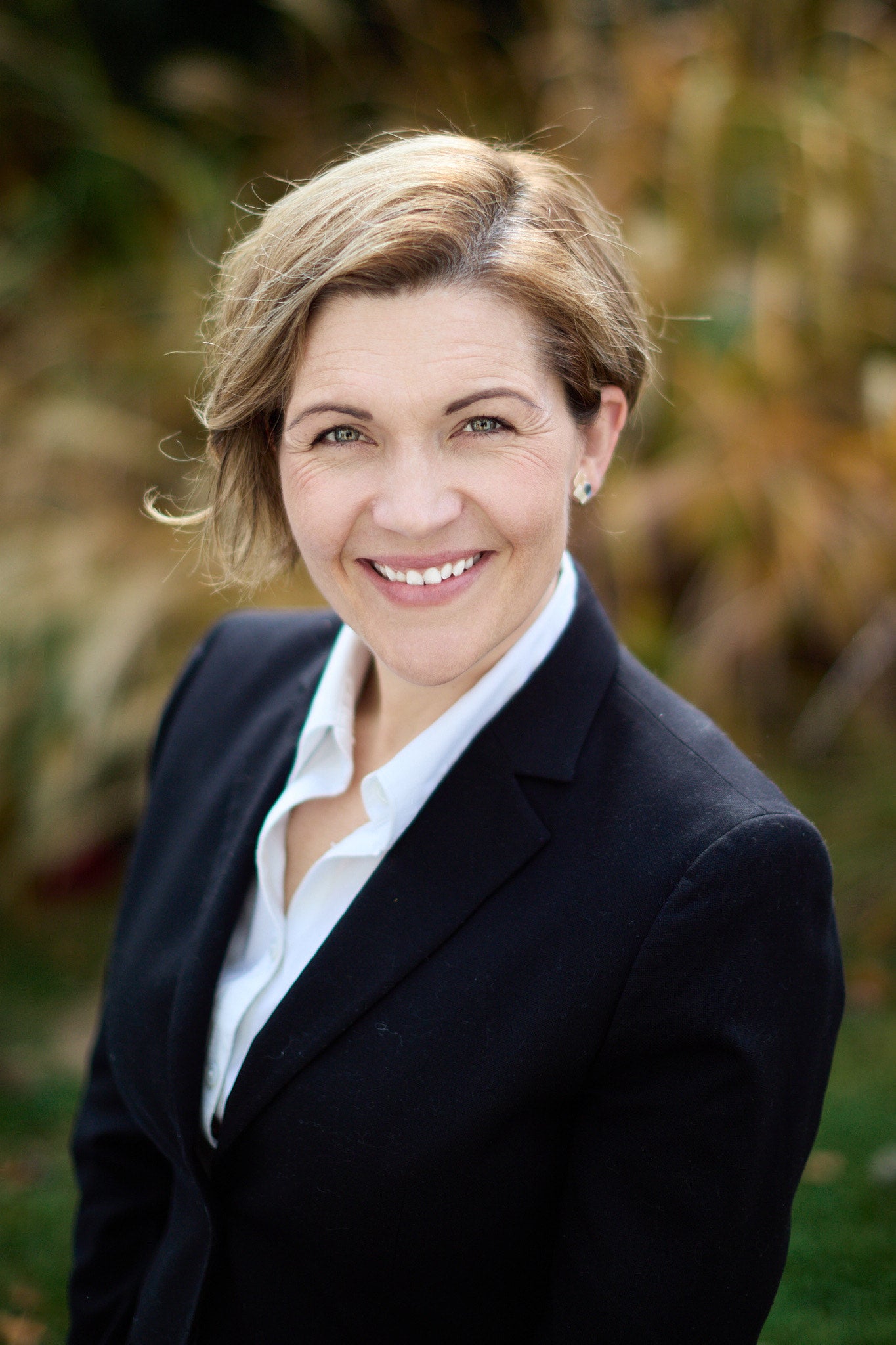Portrait of a smiling individual in a business suit, standing outdoors with blurred greenery in the background.