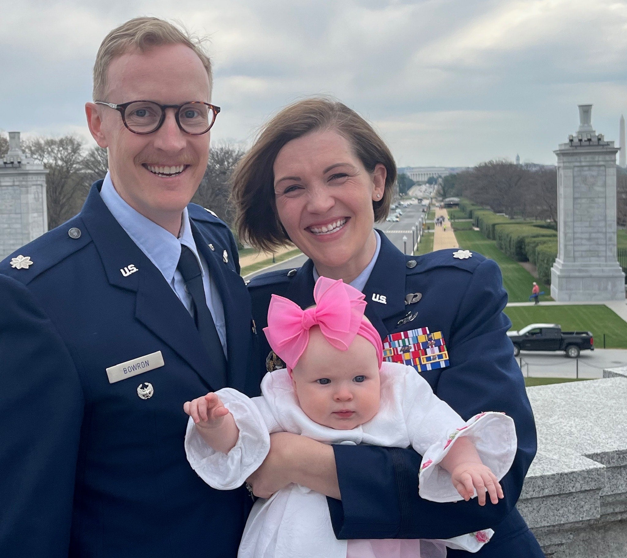 Two U.S. Air Force officers holding a baby, standing in front of Arlington National Cemetery's Memorial Amphitheater.