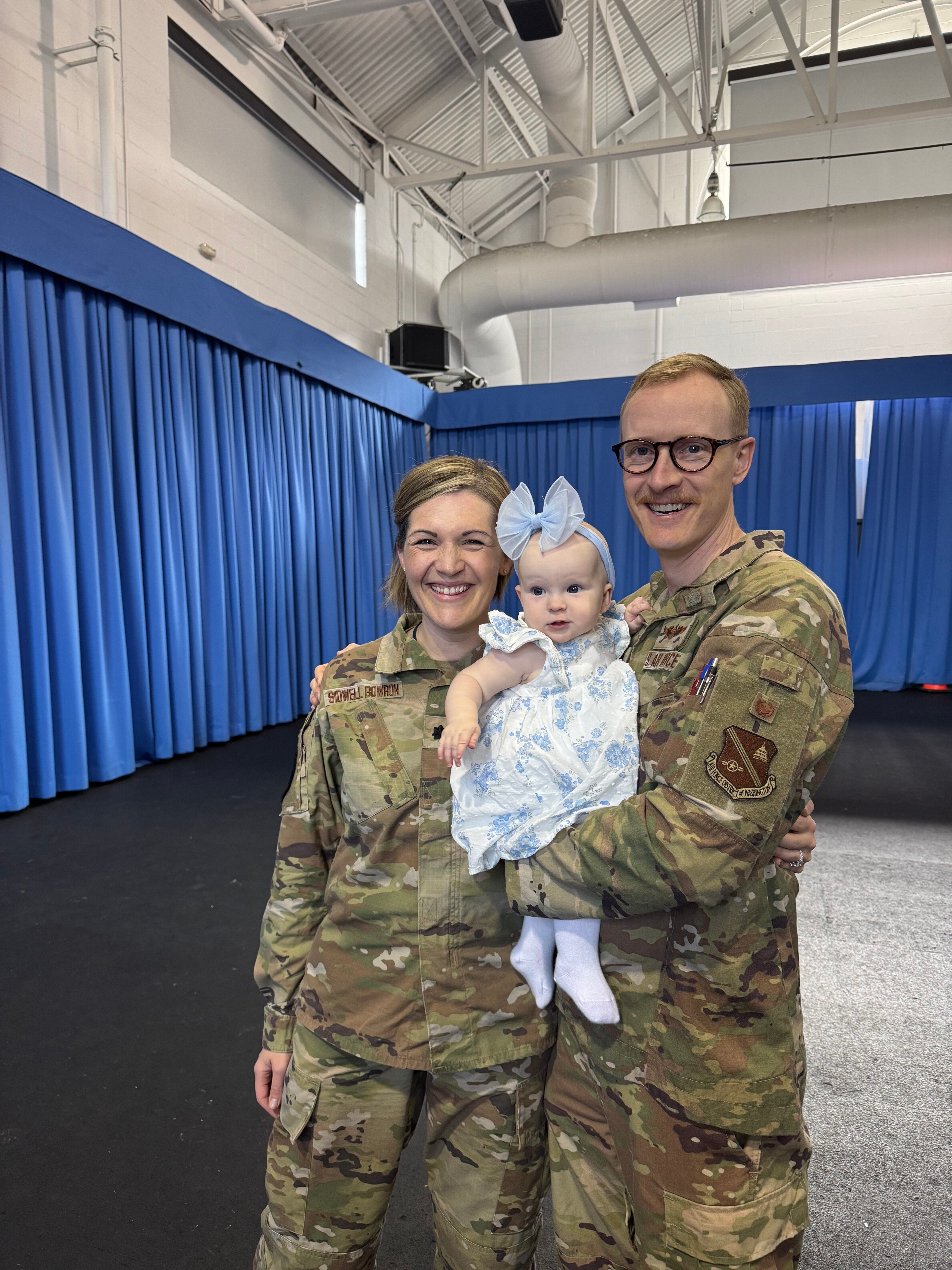 Two U.S. soldiers in camouflage uniforms smiling and holding a baby dressed in a blue outfit. They are standing indoors with blue curtains in the background.