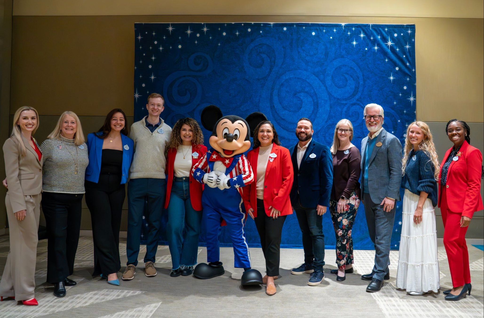 Group of people smiling at an event, standing with Mickey Mouse in front of a blue starry backdrop.