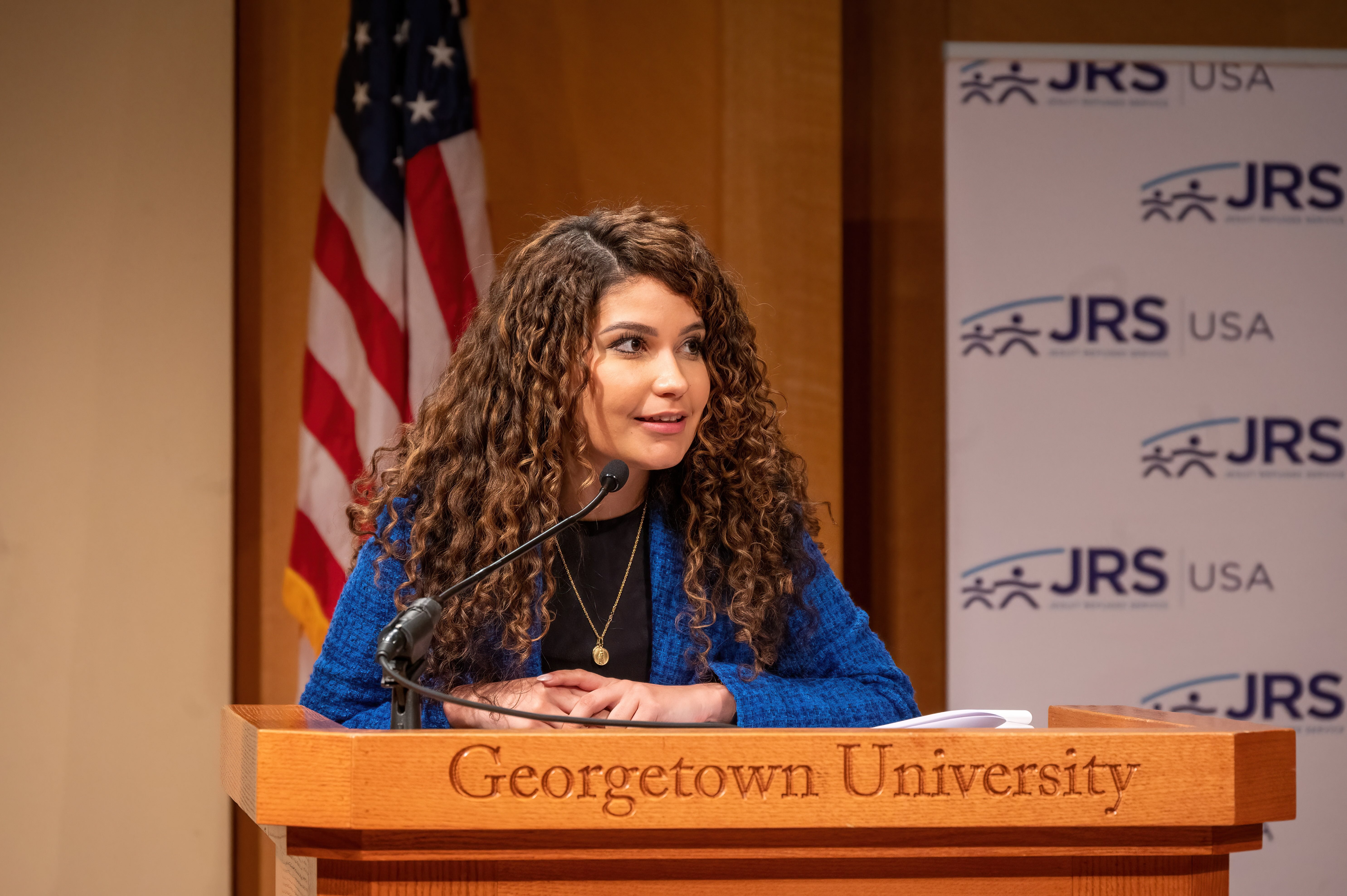 A person speaking at a podium with the Georgetown University and JRS USA logos, in front of an American flag.