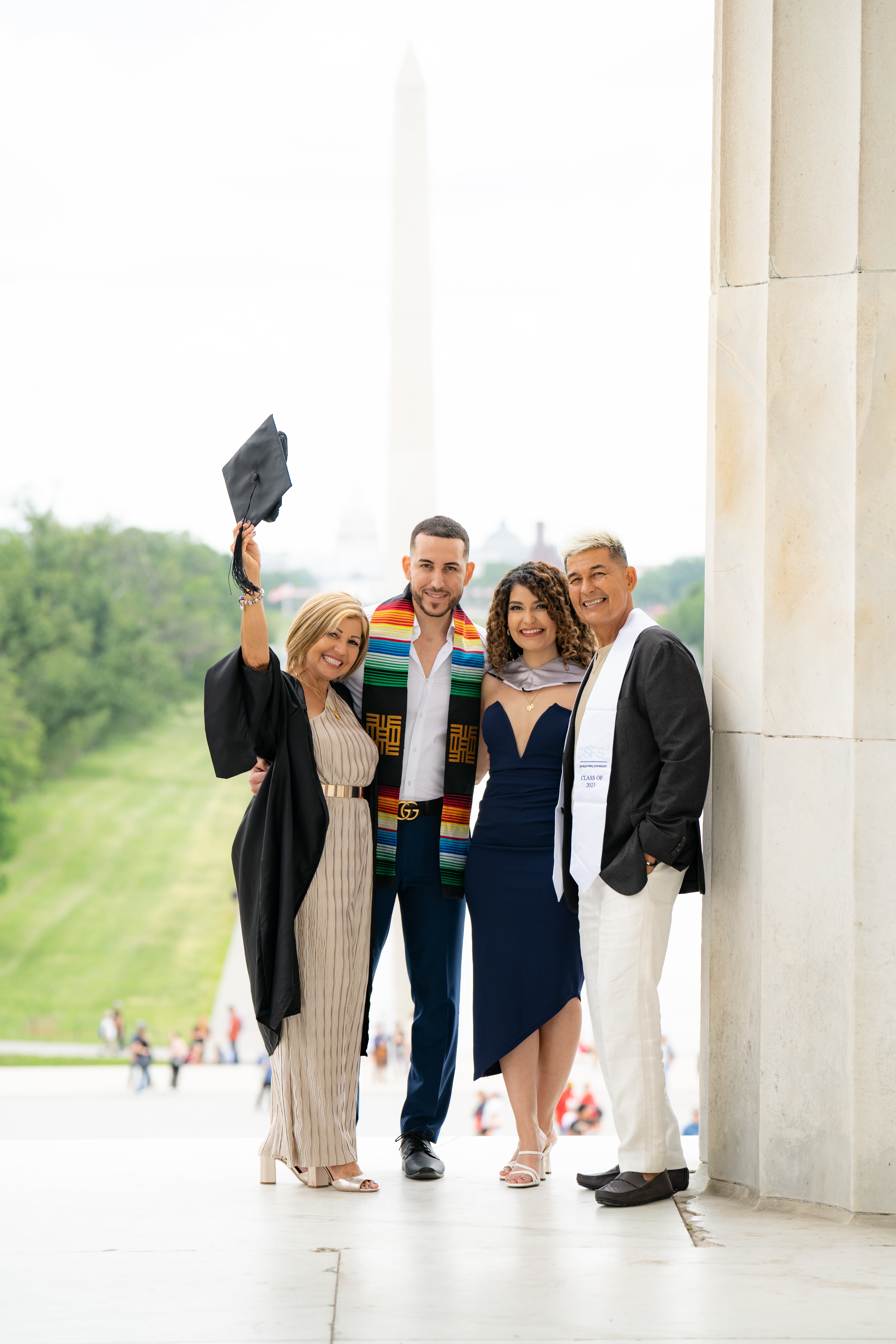 Four people celebrating a graduation, with one holding a graduation cap aloft, standing at the Lincoln Memorial with the Washington Monument in the background.