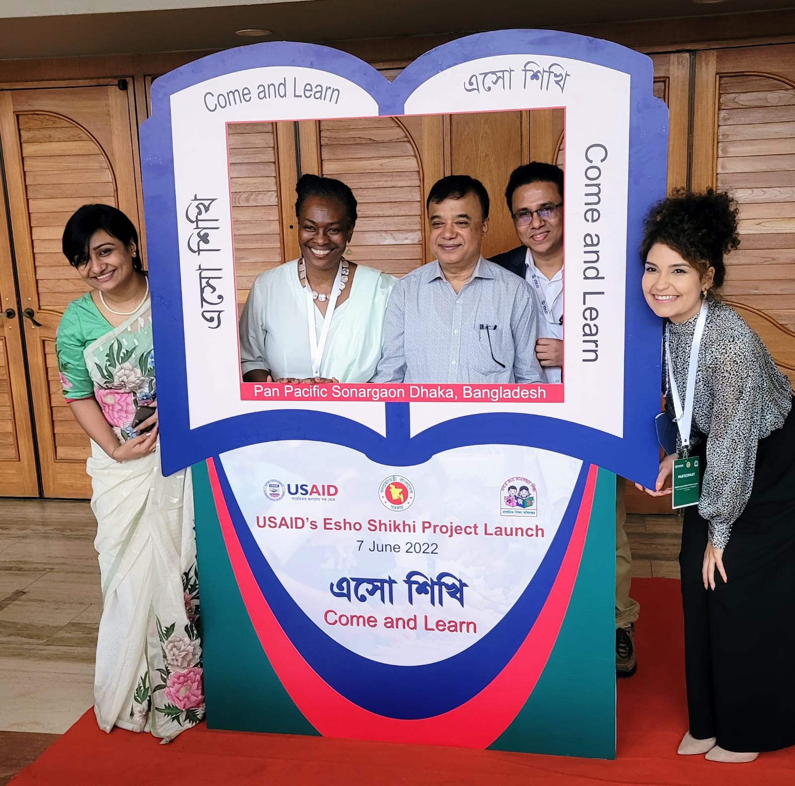 Group of five people posing with smiles at the USAID's Esho Shikhi Project Launch on June 7, 2022, at Pan Pacific Sonargaon, Dhaka, Bangladesh. They stand behind a decorative stand featuring bilingual text and the project's details.