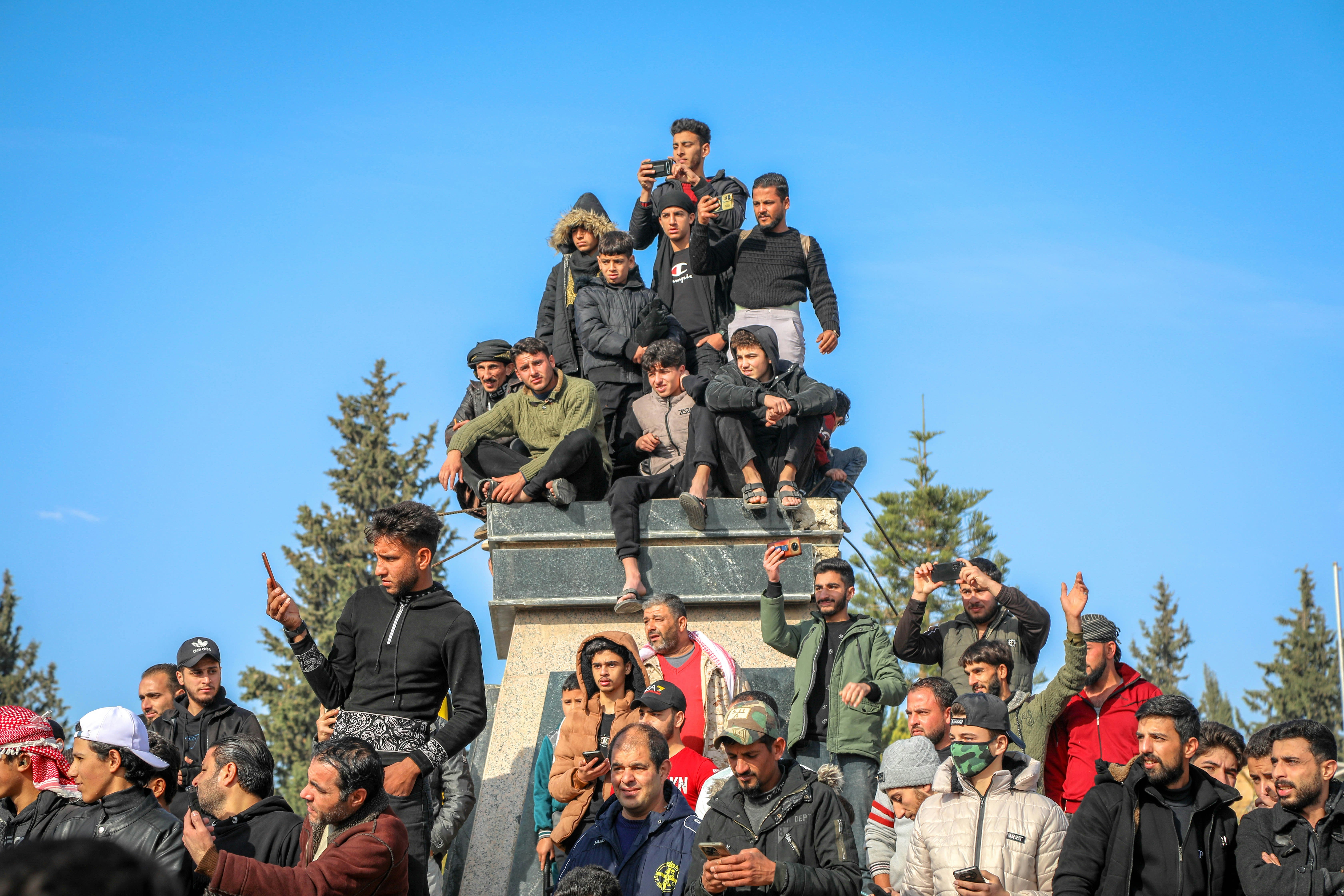 A large group of men and boys stands and crouches on and around a stone pedestal. Some hold camera phones up, taking photos.
