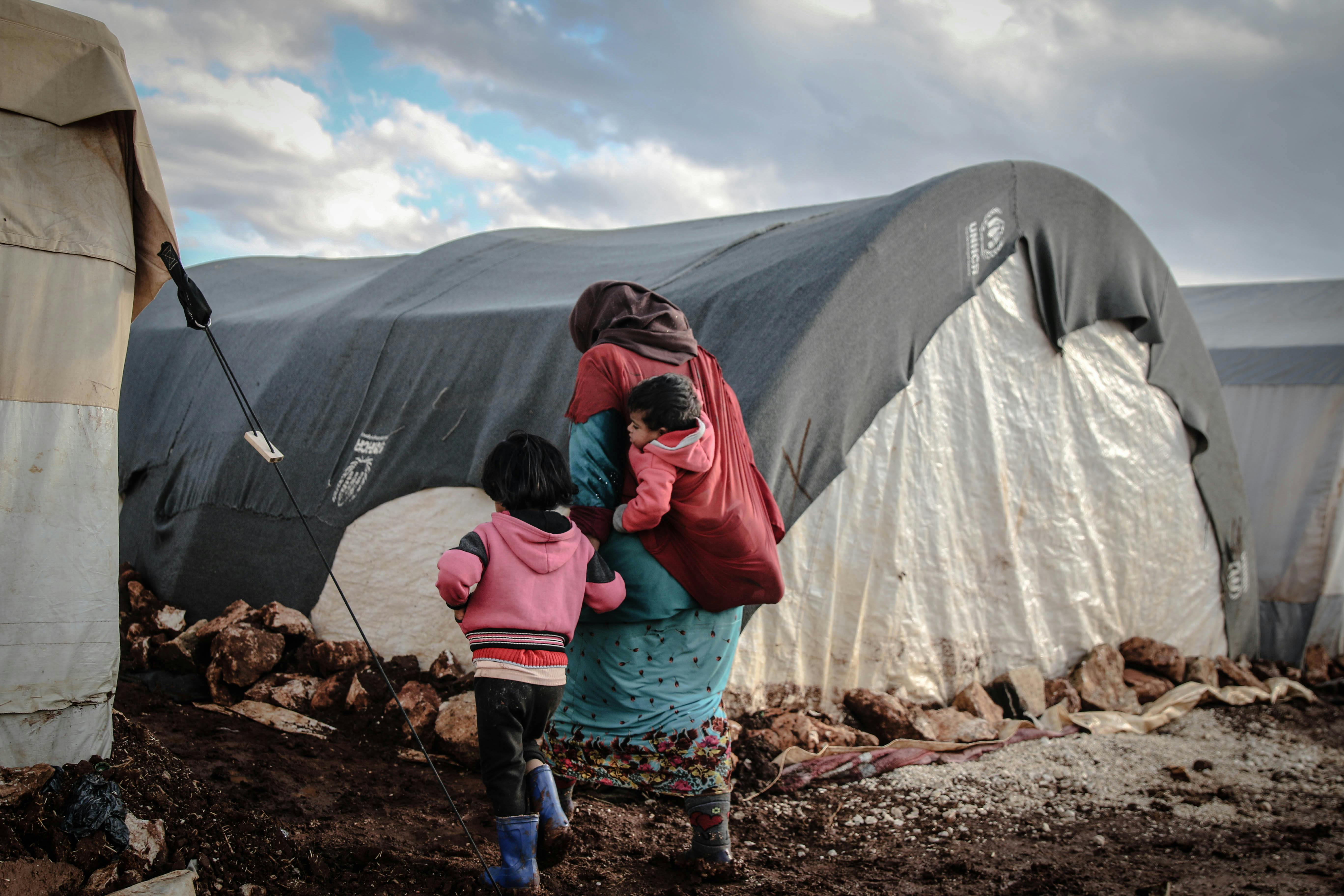 A woman with a child on her back holds hands with a small child wearing a pink jacket. They walk in mud in front of a row of white tents.