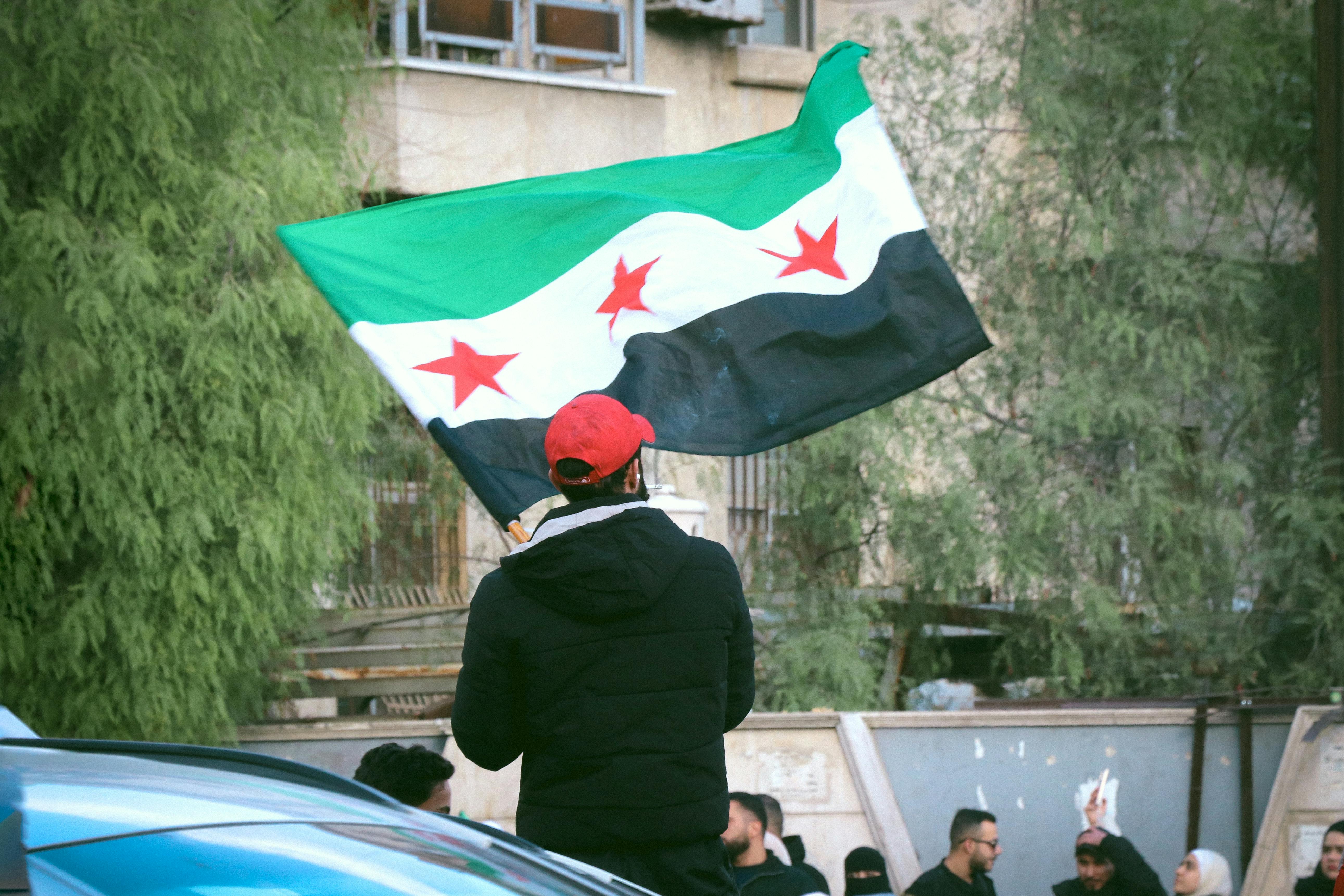 A man with a red cap and a black coat waves a Syrian flag with his back to the camera.