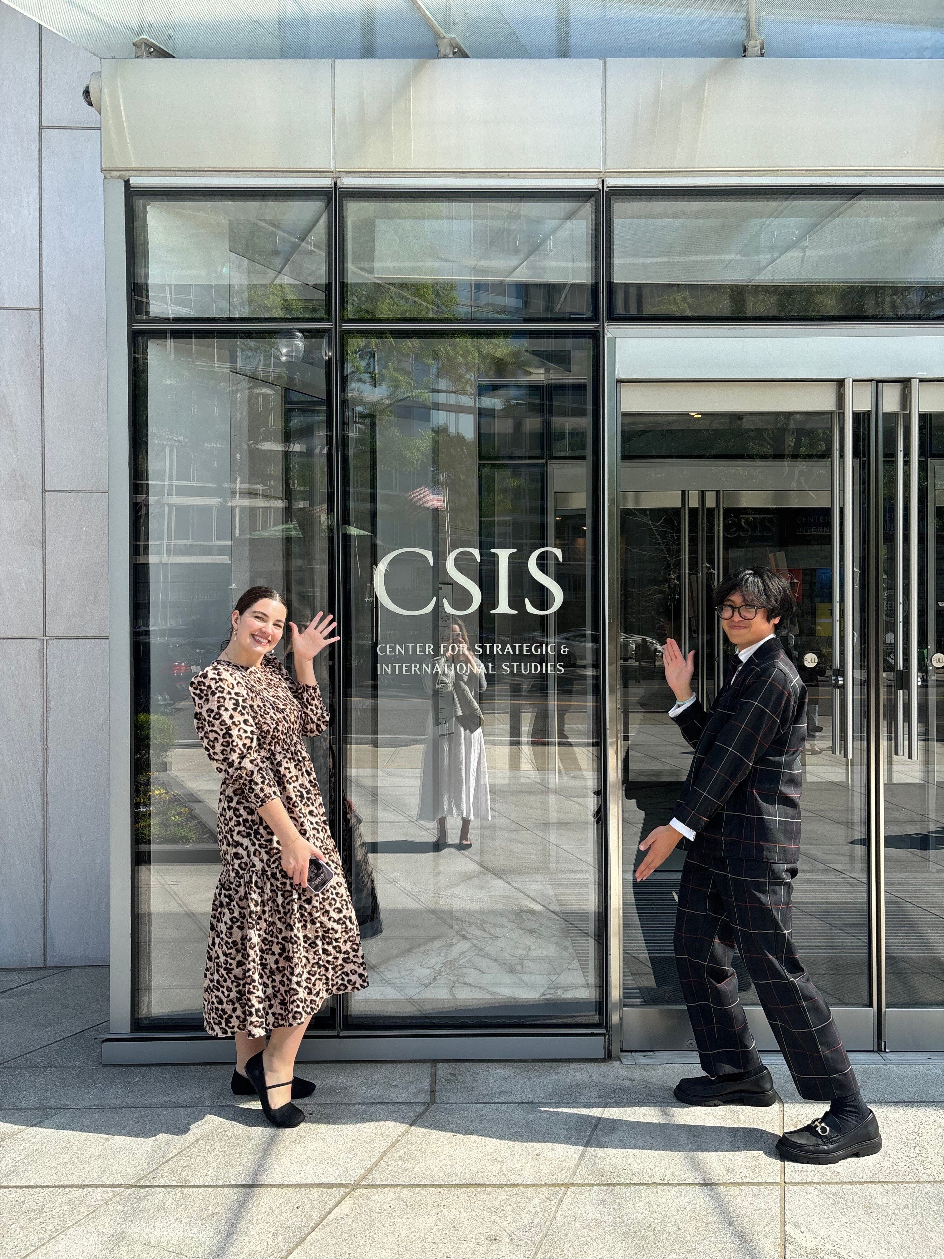 Two individuals playfully posing outside a building with glass doors labeled 'CSIS.' One is standing and waving, dressed in a pinstripe suit, and the other is posing with one leg raised, wearing a leopard print dress.