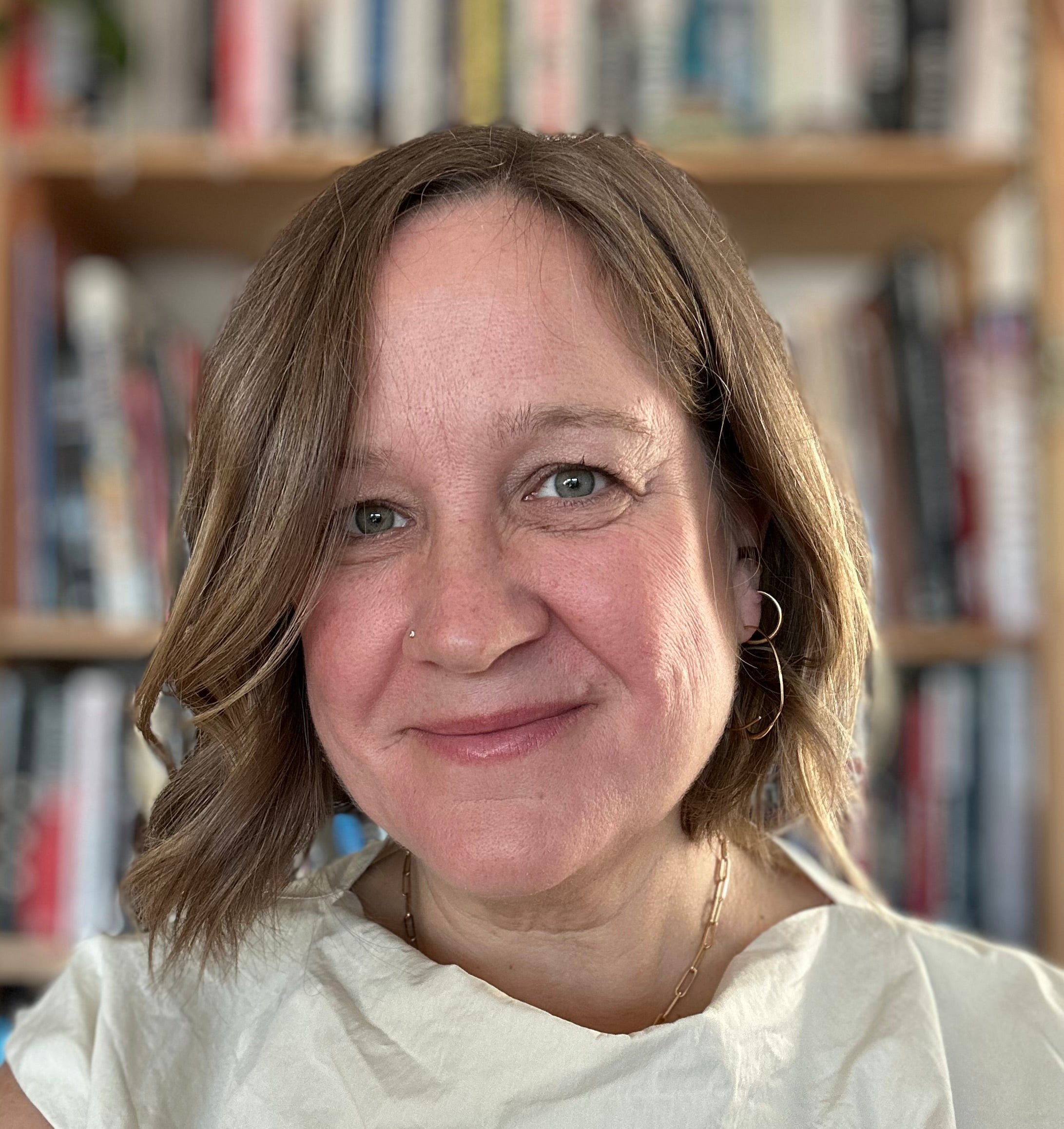 A woman with short blonde hair wearing a white top and gold chain necklace. A bookcase is blurred in the background.
