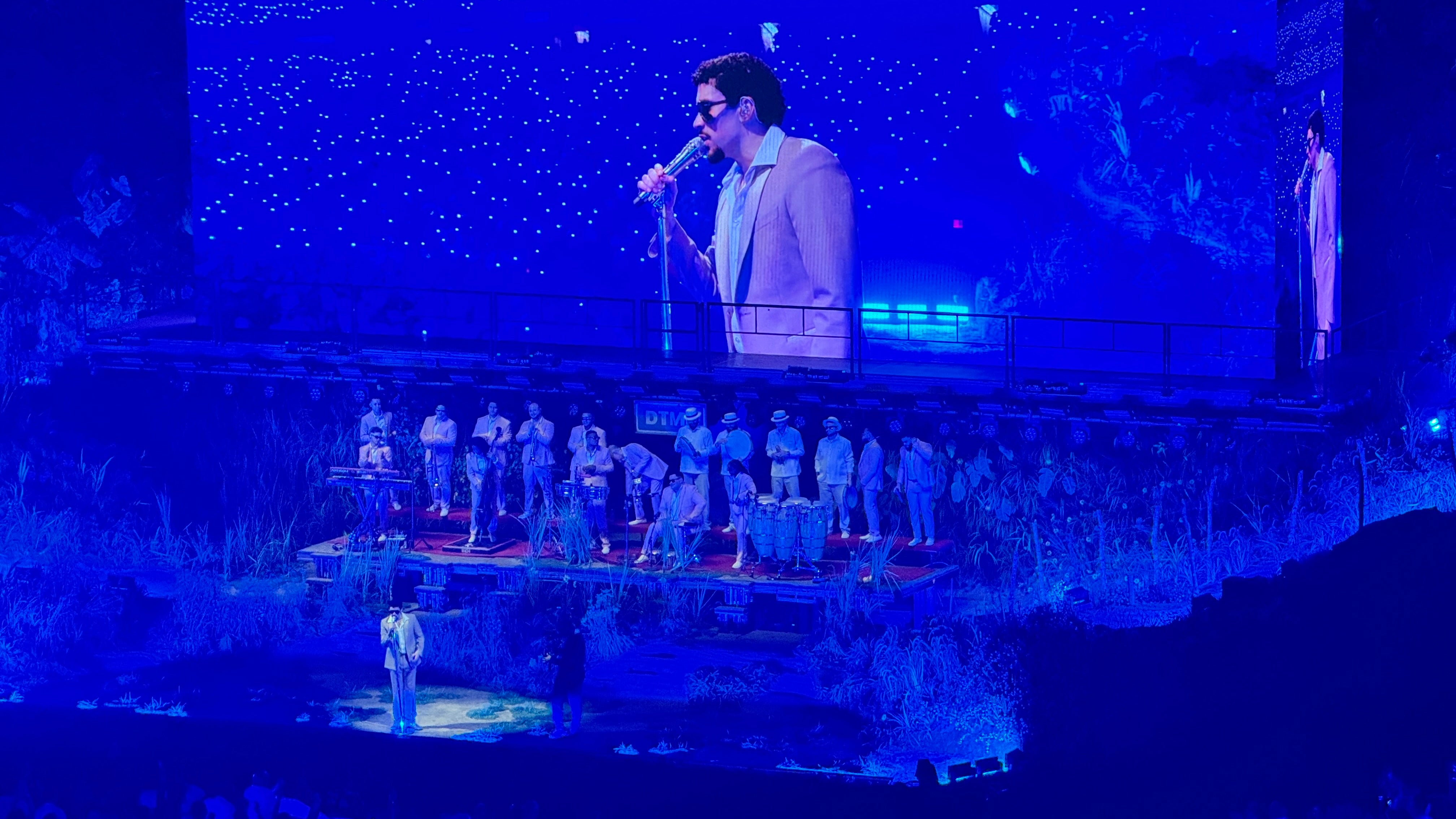 Bad Bunny stands on a stage with a band behind him and his image projected on the screen behind him.