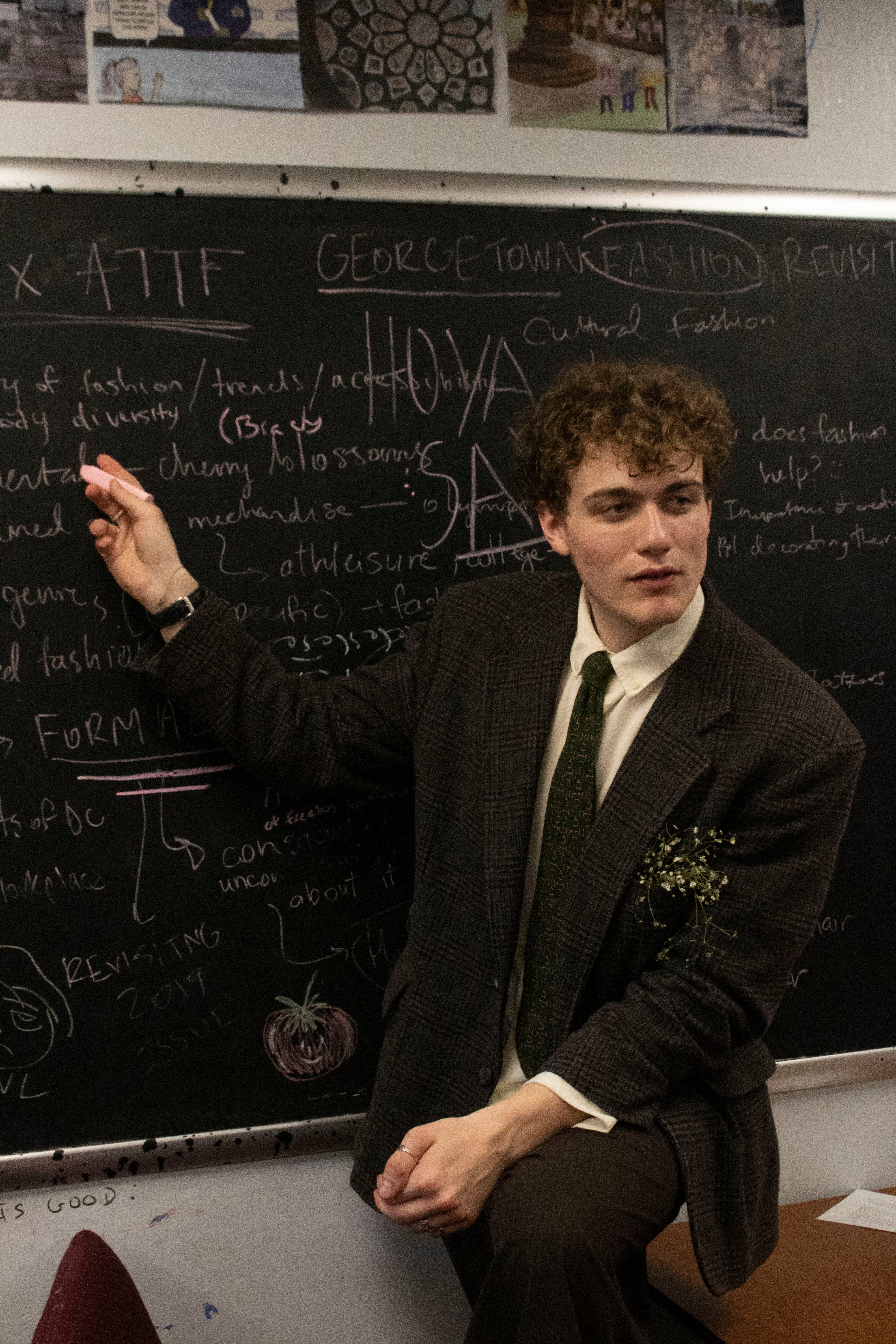 A person in formal attire stands in front of a chalkboard covered in handwritten text and diagrams related to fashion. The person is holding a piece of chalk and gesturing towards the board. "Georgetown" and "Hoya" are prominently written on the board.