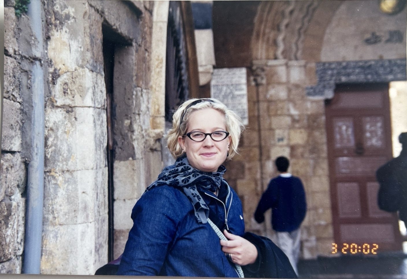 A woman with short blonde hair with glasses wearing a blue jacket and scarf in Lebanon.