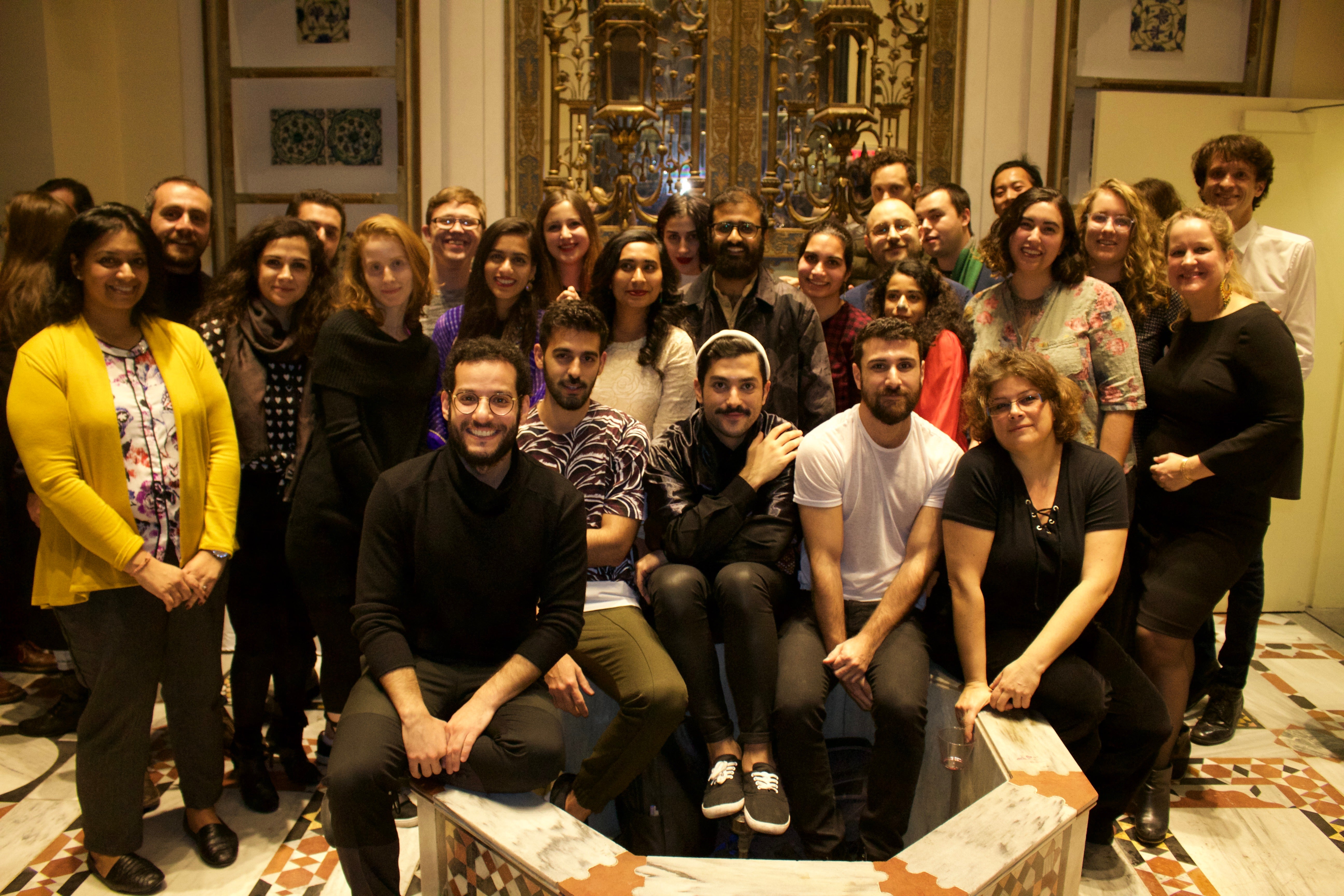 A group of individuals pose for a photo in front of an ornate decorative door. The setting appears indoors, with patterned flooring visible. The group consists of around 25 people, arranged in rows, with some standing and others sitting on a marble structure in the front.