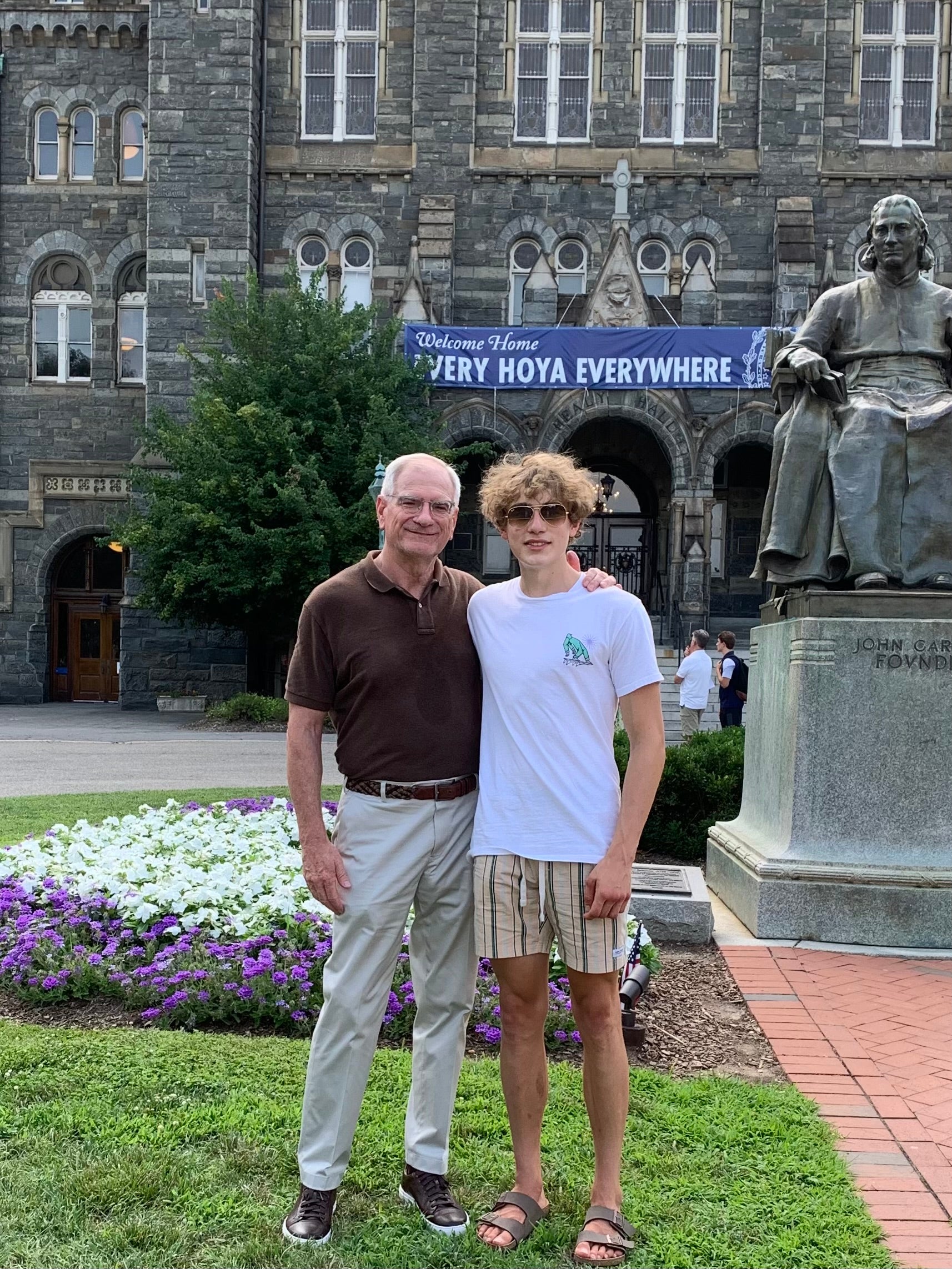 Two people stand together smiling in front of a historic building with Gothic architecture. A statue of John Carroll is nearby. They are standing on a grassy area with purple and white flowers.