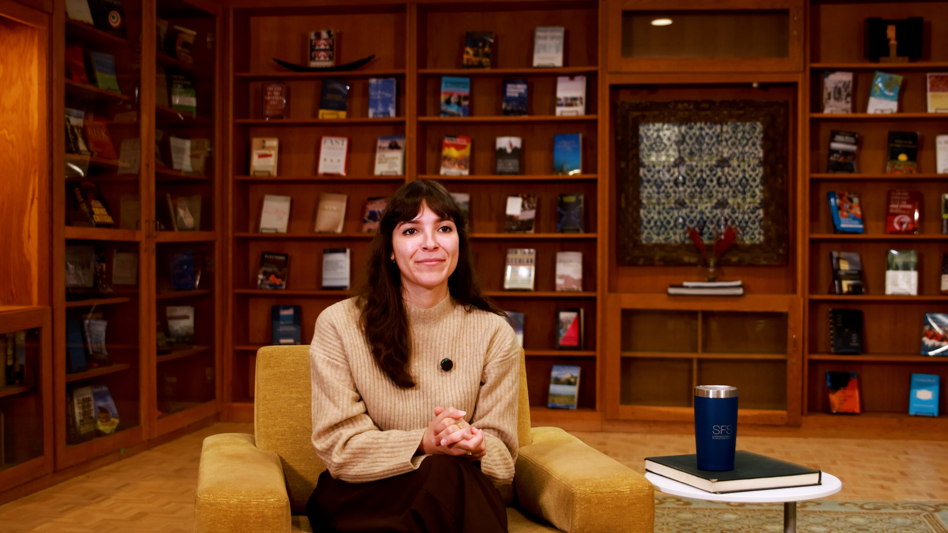 A person sits in a cozy library setting with wooden bookshelves filled with various books and decorations in the background. They are seated on a cushioned chair next to a small table holding a blue SFS-branded tumbler.