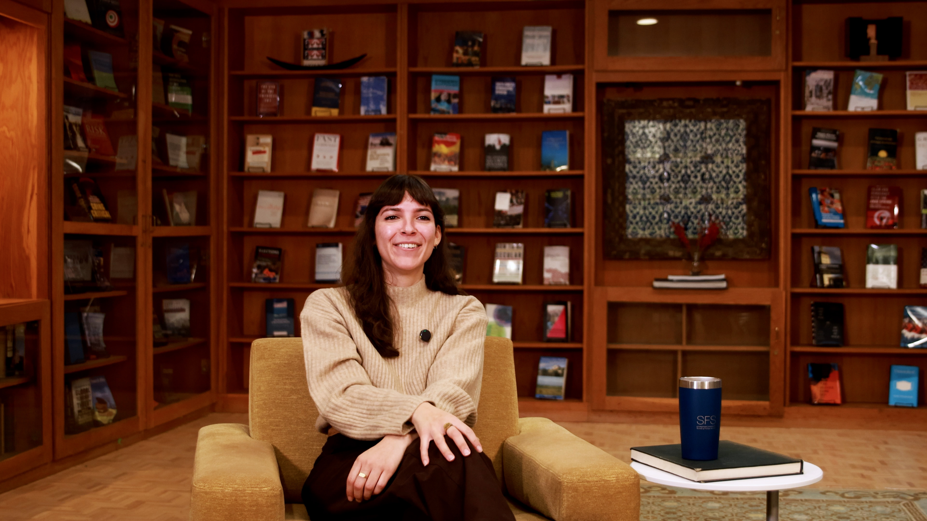 A person sits in a cozy library setting with wooden bookshelves filled with various books and decorations in the background. They are seated on a cushioned chair next to a small table holding a blue SFS-branded tumbler.