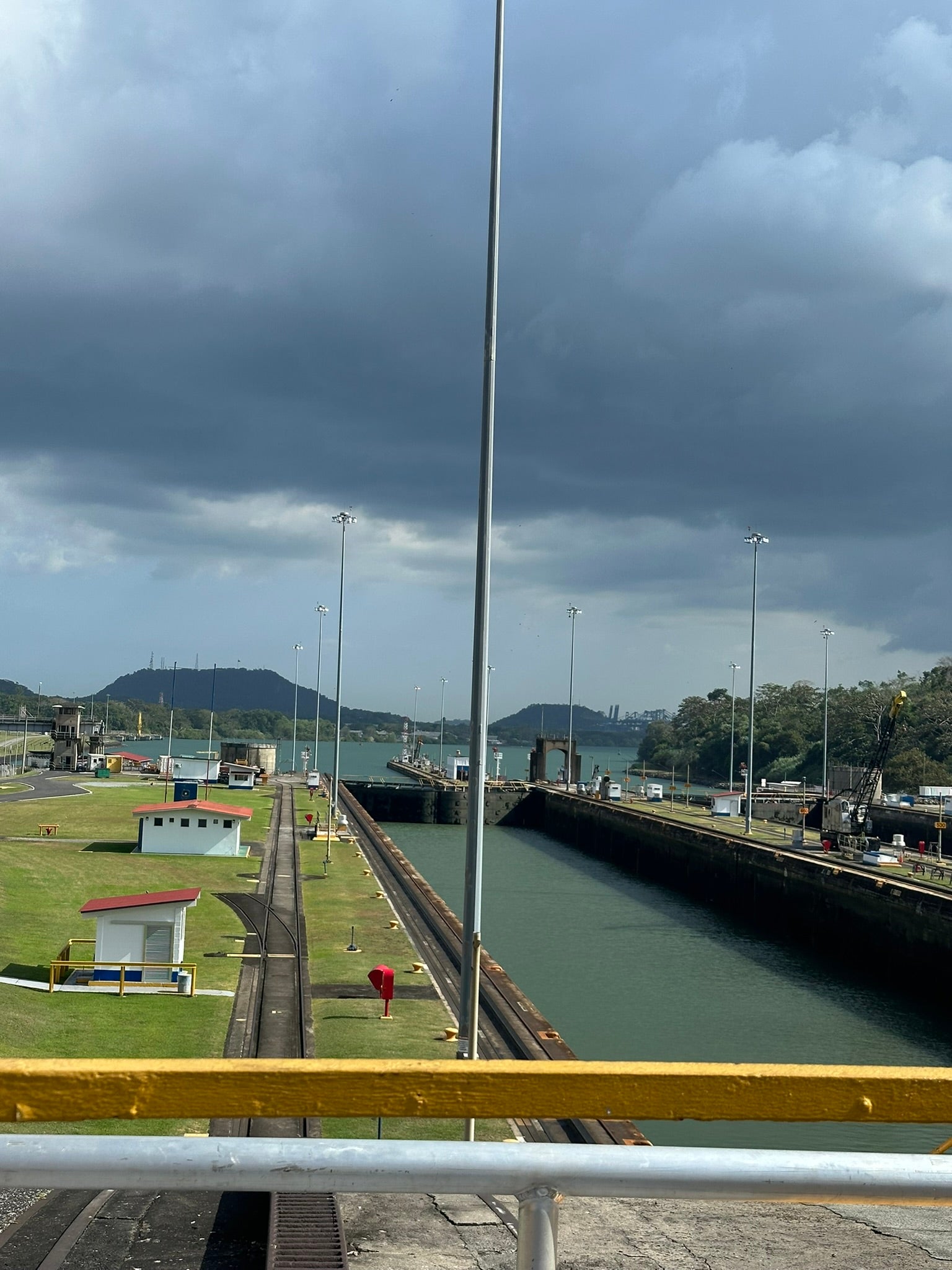 View of the Panama Canal with shipping lanes and gates visible, flanked by grassy areas and maintenance buildings. Dark clouds loom overhead, with lush green hills in the background.