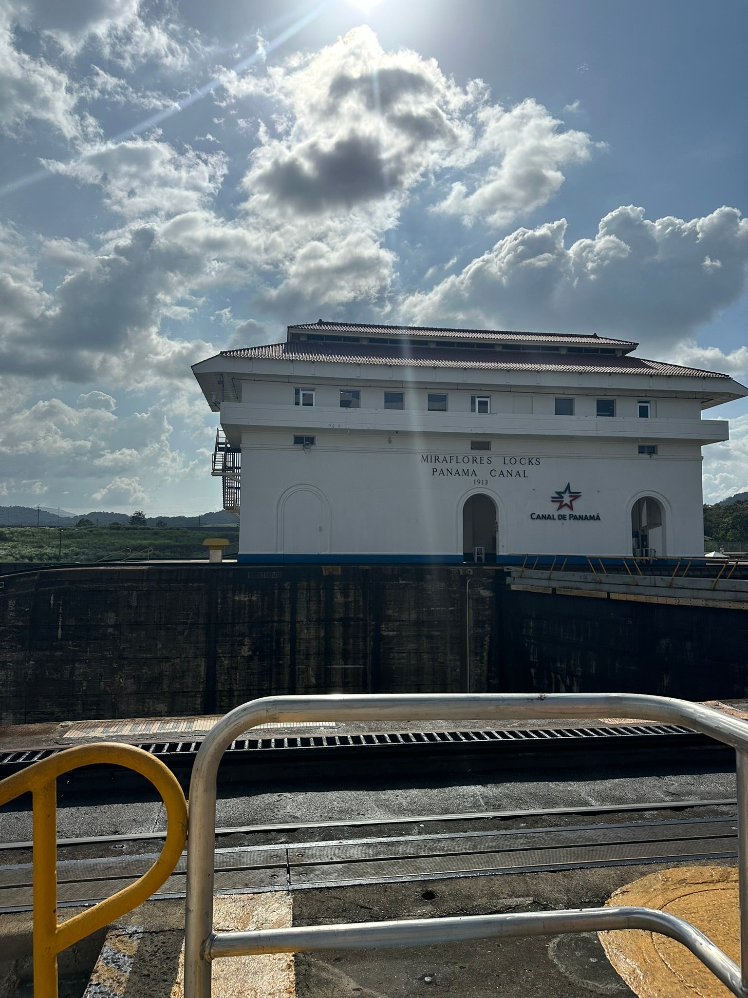 View of the Miraflores Locks building at the Panama Canal. The sun is shining, illuminating the structure with a partly cloudy sky in the background. A railing is visible in the foreground.