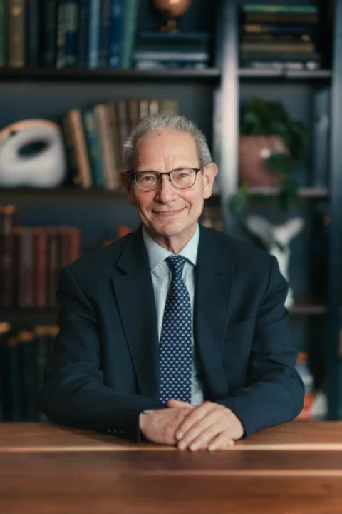 A man with glasses, grey hair, and a dark blue suit with a blue necktie.