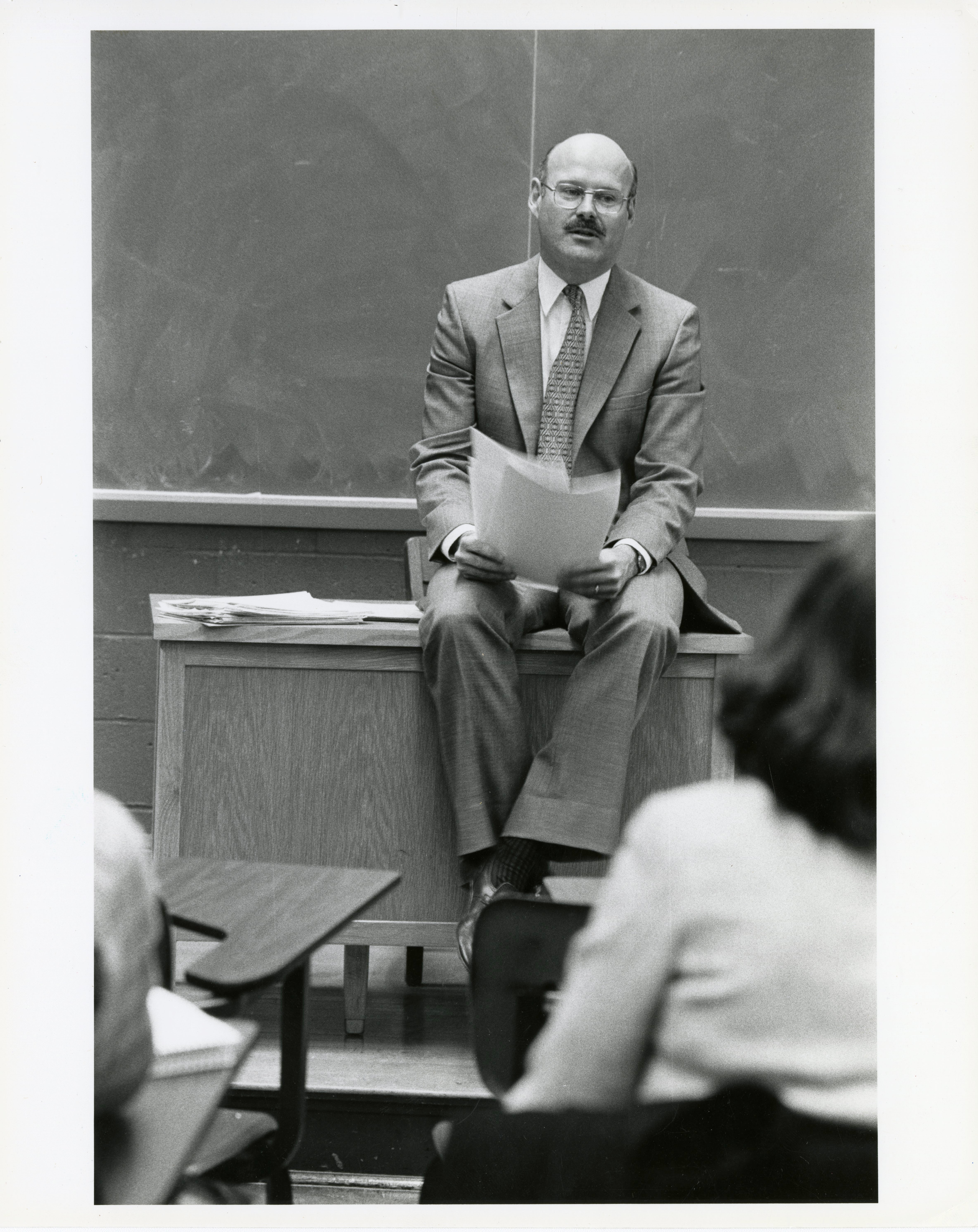 Michael Hudson sits on a desk in front of a chalkboard wearing a suit.