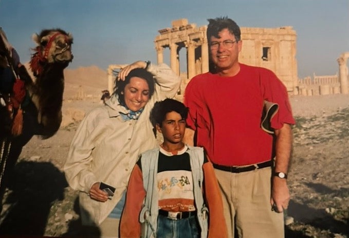 Two adults and a child stand in front of an archeological ruin. A camel is also in the frame on the left.