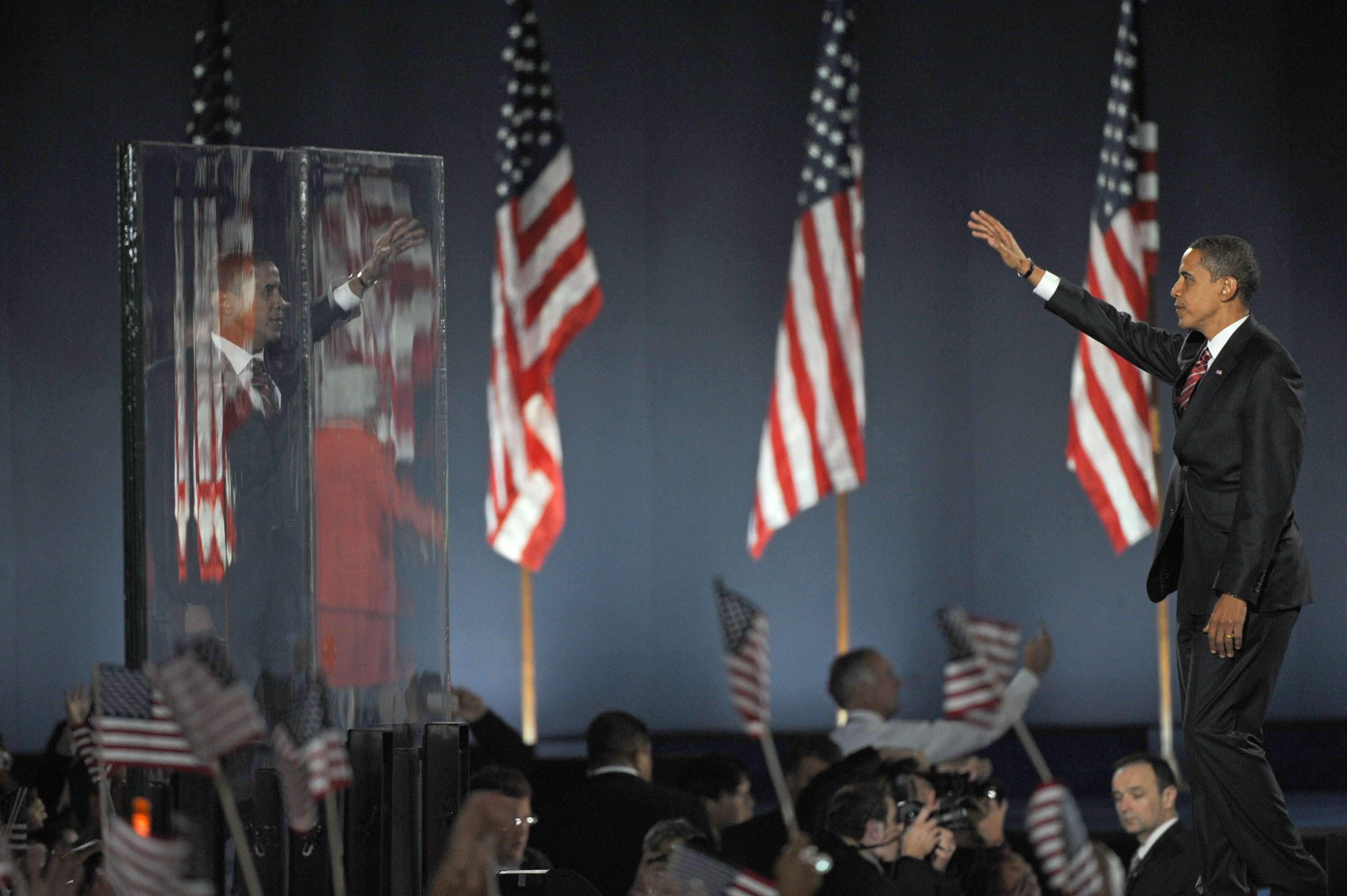 Barack Obama on stage waves to a cheering crowd while being reflected in a clear barrier. Numerous American flags are visible in the background.