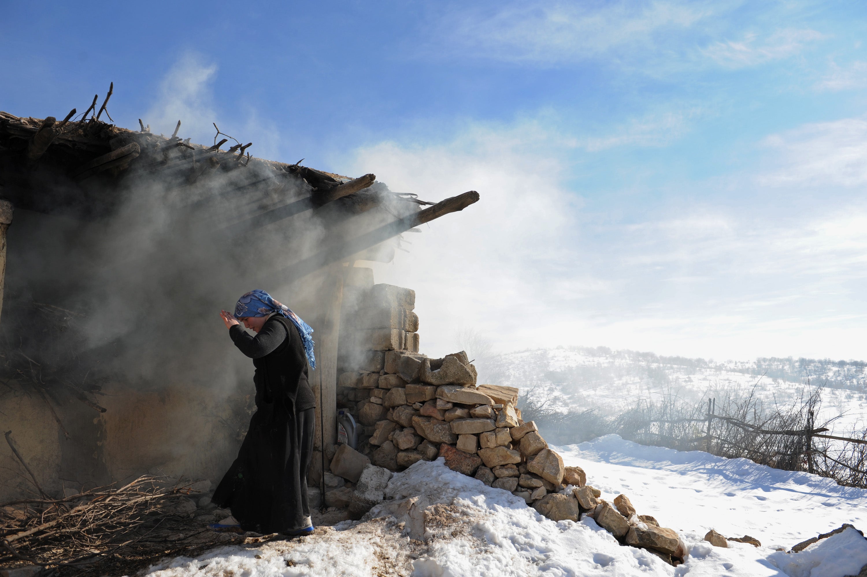 A person stands near a rustic, smoking structure made of wood and stone, set in a snowy landscape under a clear blue sky. They appear to be shielding their face from the smoke. A scenic view of snow-covered hills is visible in the background.
