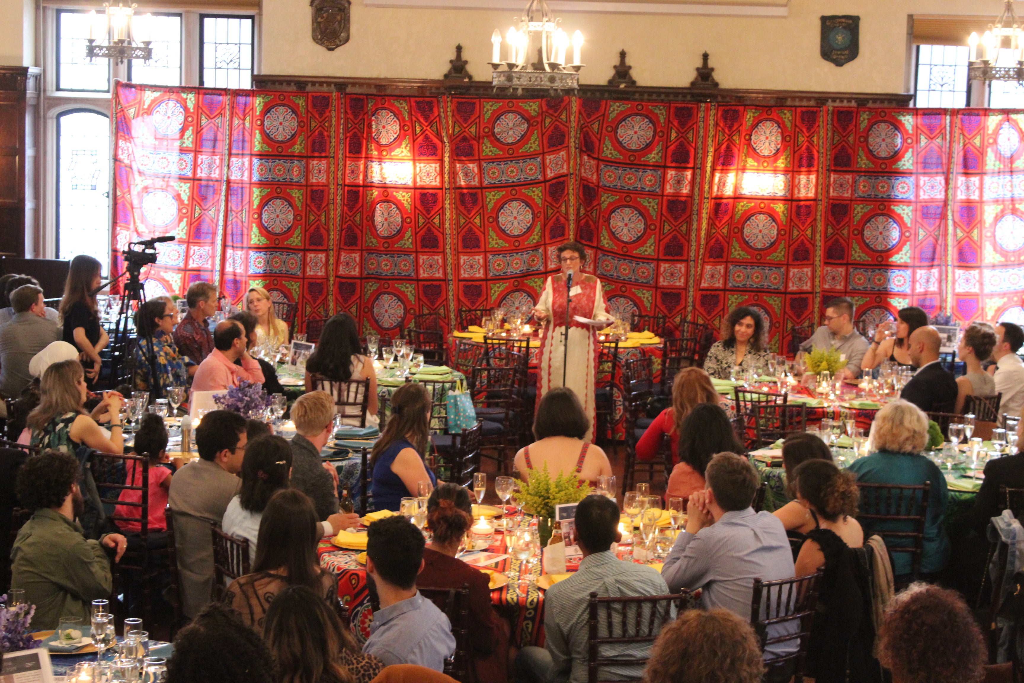 Fida Adely speaks at a microphone in front of a bright colored tapestry.