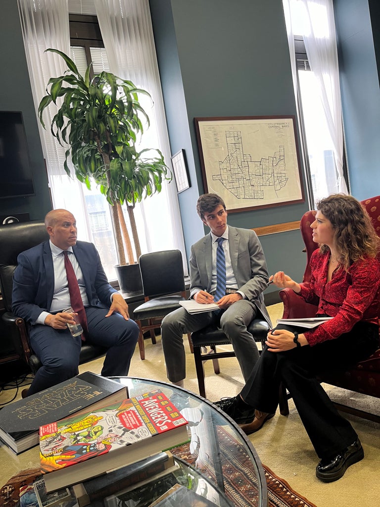 Three people sit and converse in an office setting with large windows. One person gestures while speaking. A tall plant and a large map adorn the room. Magazines about superheroes are visible on a glass table.