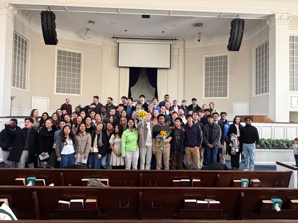 A large group of people poses together inside a spacious auditorium with a high ceiling and large windows. Two individuals in the front are holding bouquets of sunflowers. The room has a central stage with an empty screen and pew-style seating.