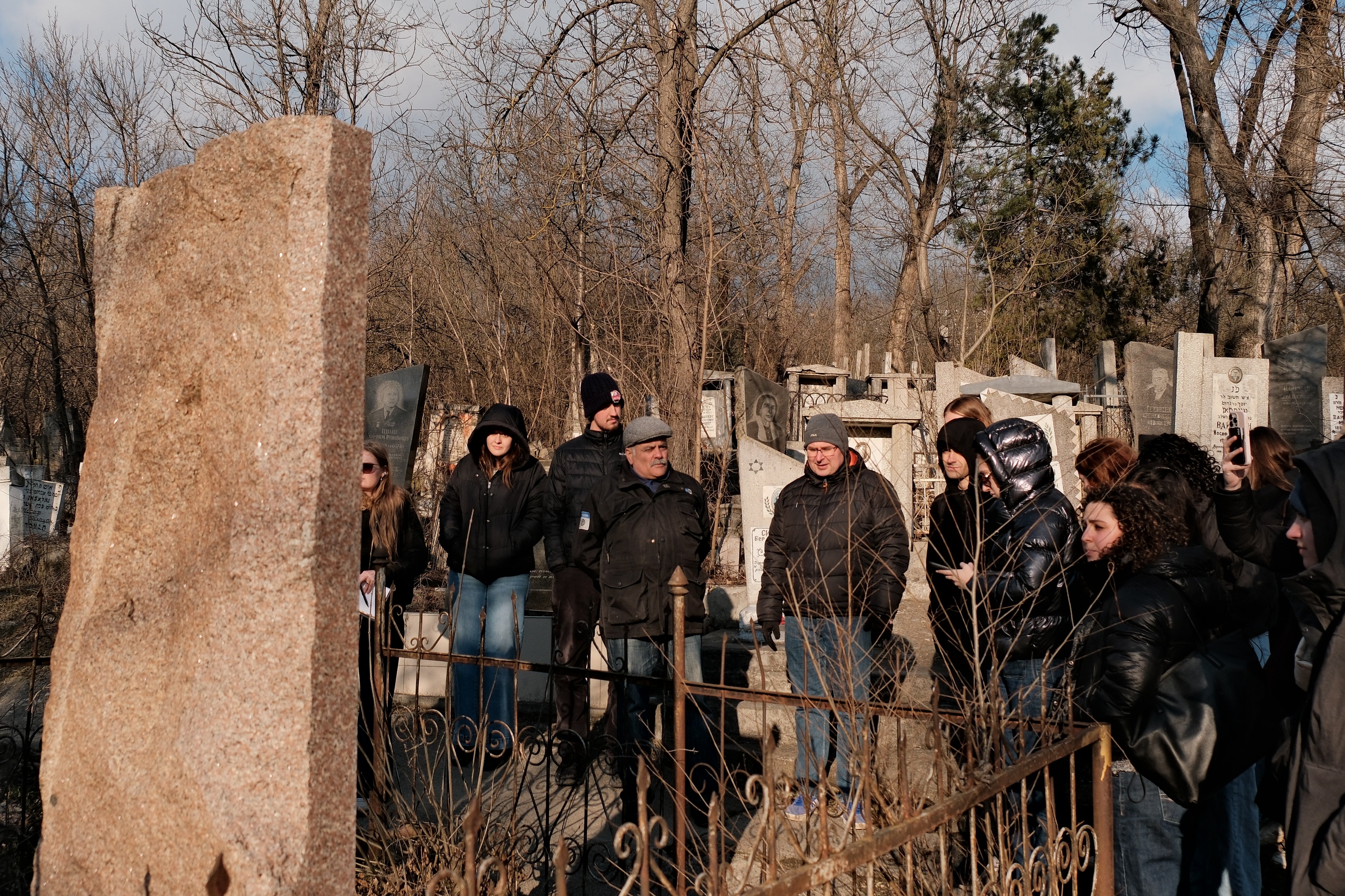 A group of people stands in an outdoor setting surrounded by numerous gravestones. They are gathered near a large stone monument. Bare trees and various headstones fill the background, suggesting a cemetery. The sky is clear with some clouds.
