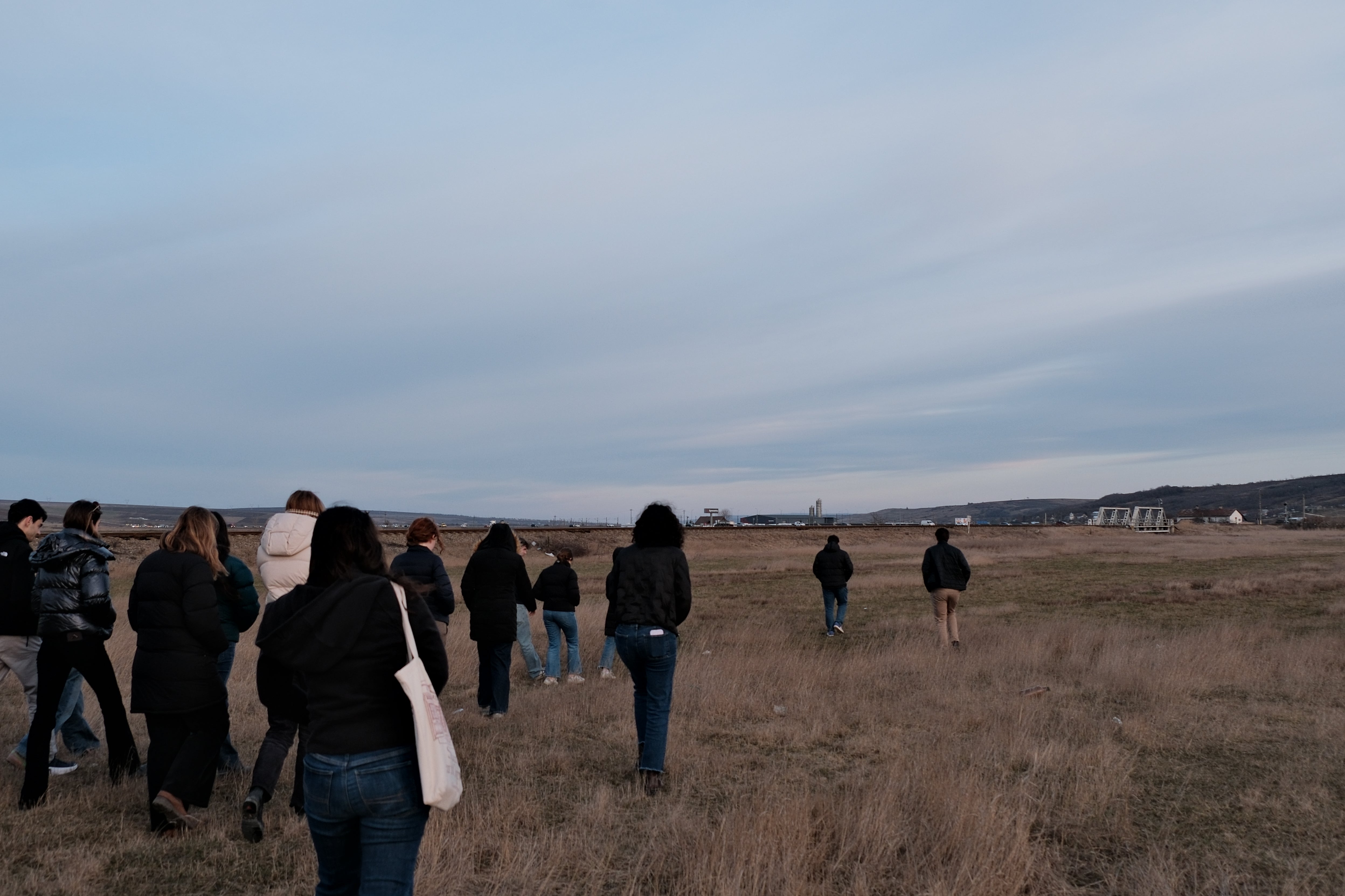 A group of people walking across a grassy field under a vast, cloudy sky. The landscape is open, with distant buildings and a horizon in the background.