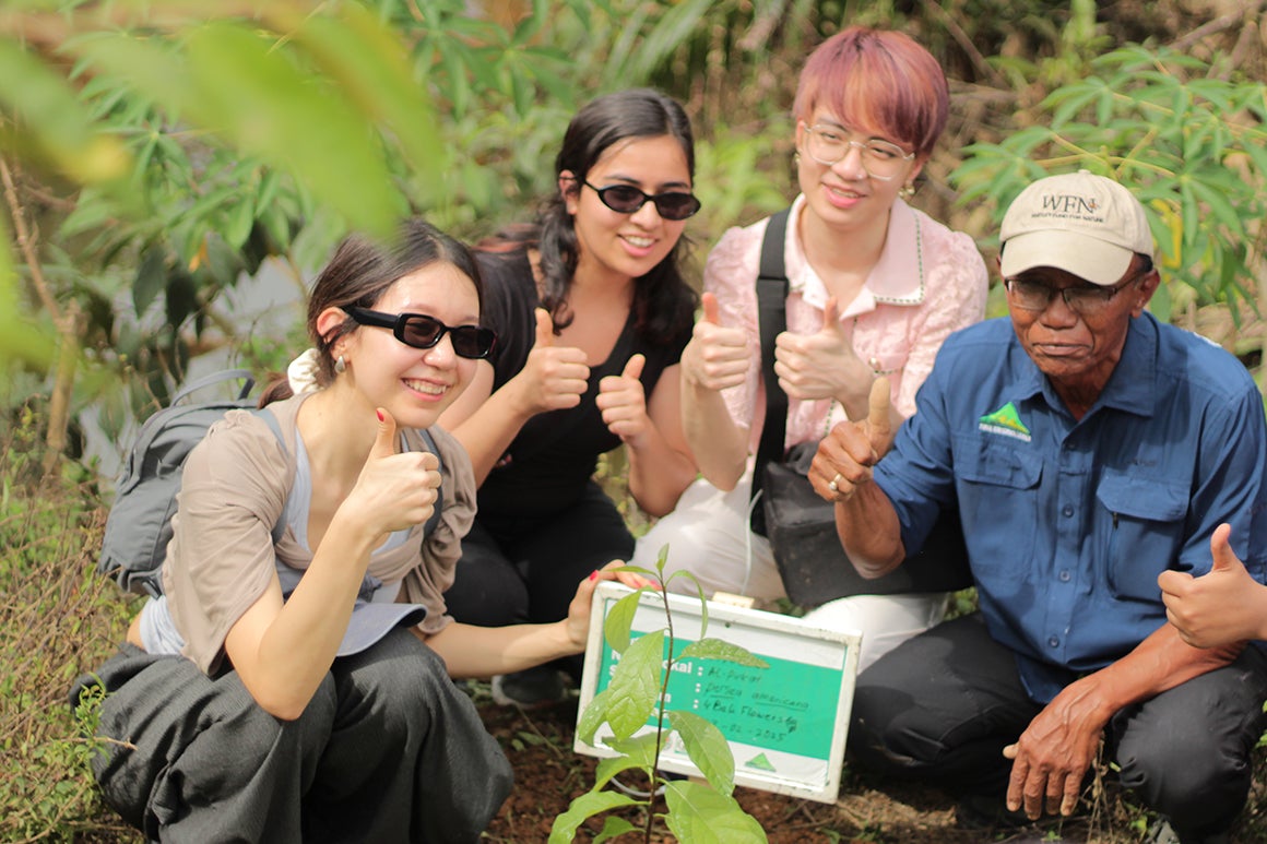 three young women and an older man crouching in nature in front of a small plant with their thumbs up