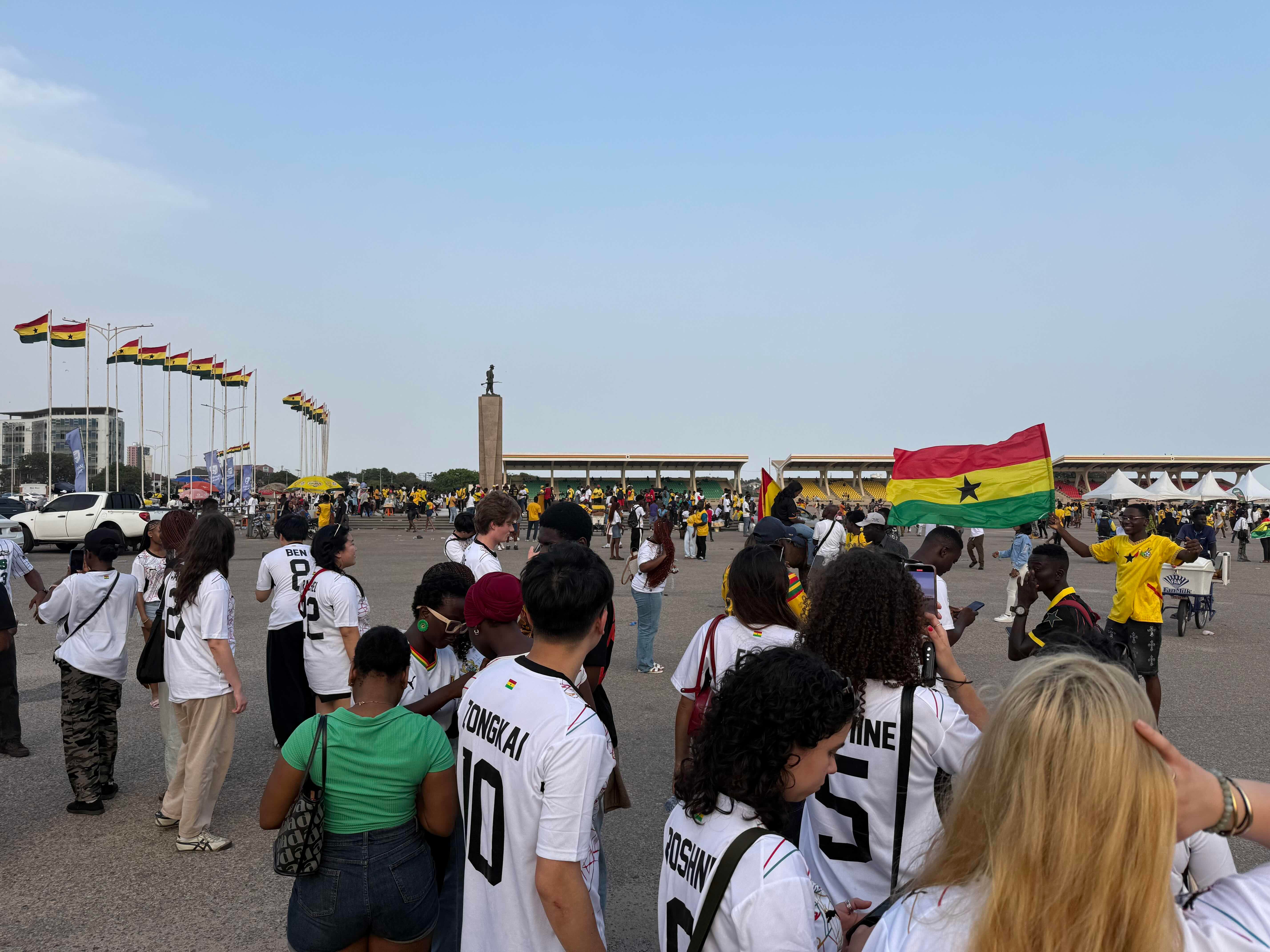 A crowd gathers in a large open area, many wearing sports jerseys. Several people hold Ghanaian flags. In the background, there are multiple flagpoles with Ghana flags and a prominent statue.