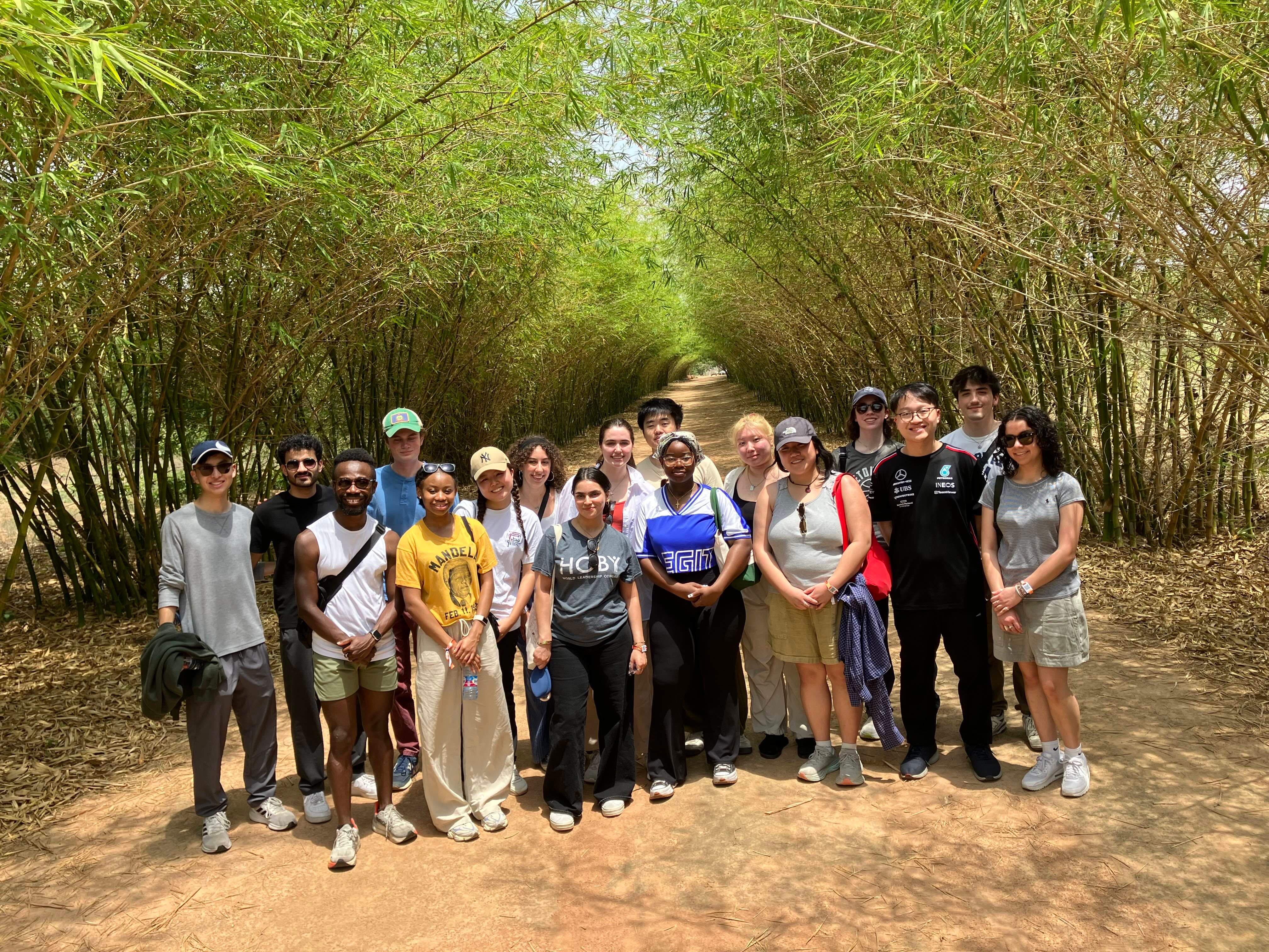 A group of people standing on a path surrounded by tall bamboo plants, smiling at the camera. The setting is outdoors, with a natural, lush environment.
