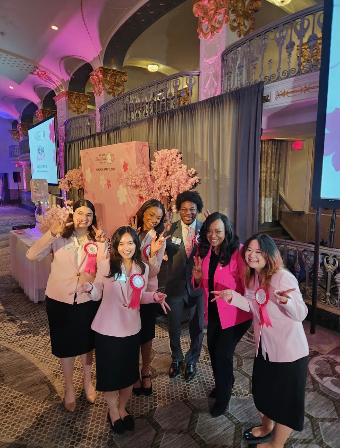 Six people pose for a group photo in an indoor venue with pink lights and cherry blossoms in the background.