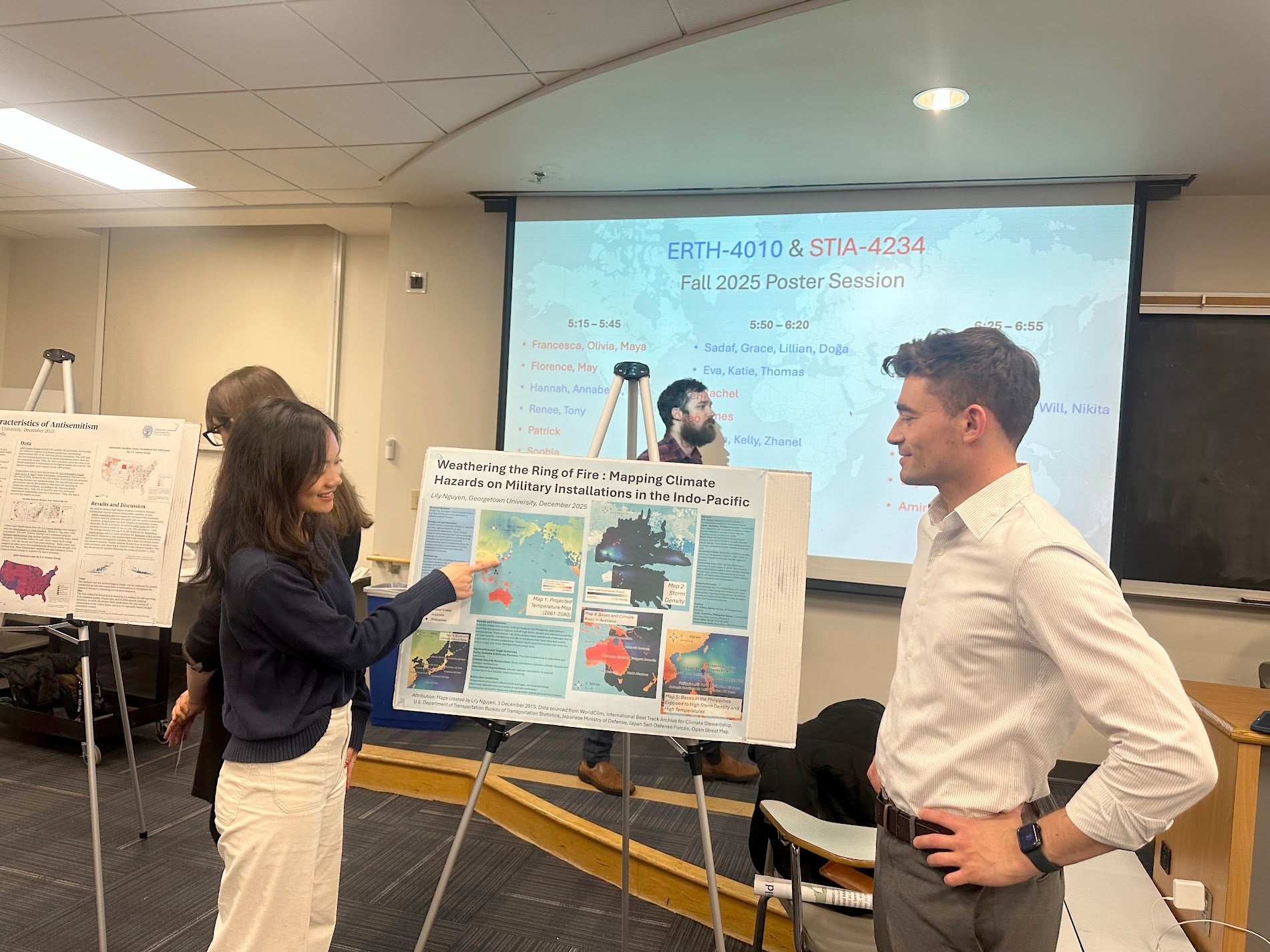 Lily Nguyen (MSFS'26) points to her poster for her final project, “Weathering the Ring of Fire: Mapping Climate Hazards on Military Installations in the Indo-Pacific." Another person appears to be listening. The setting appear to be inside, with a white screen projecting "ERTH-4010 & STIA-4234" in the background.