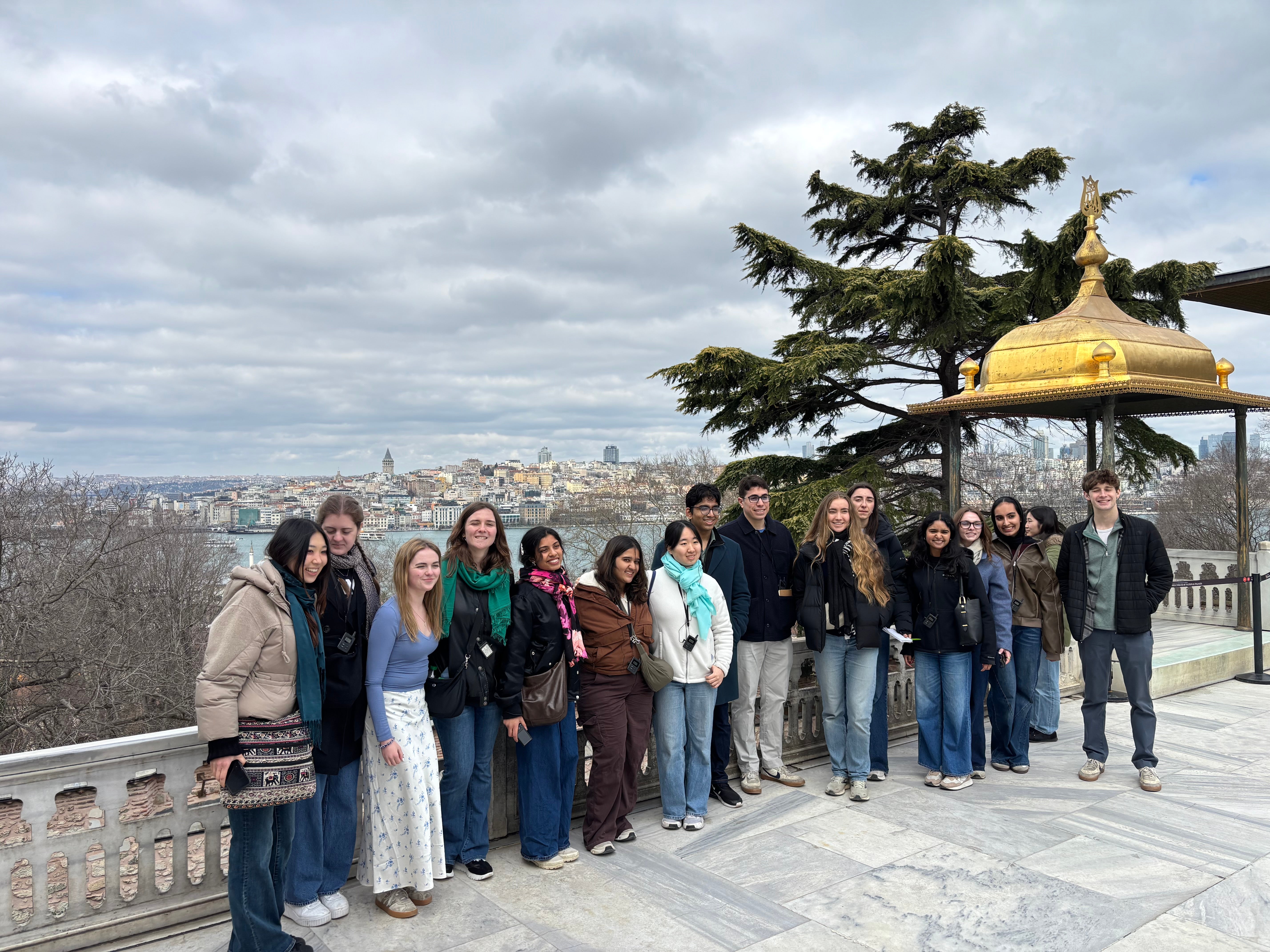 A group of people posing together on a scenic terrace with a view of Istanbul. In the background, the cityscape stretches across the horizon under a cloudy sky, and a tree stands nearby. A golden pavilion roof is visible on the right.