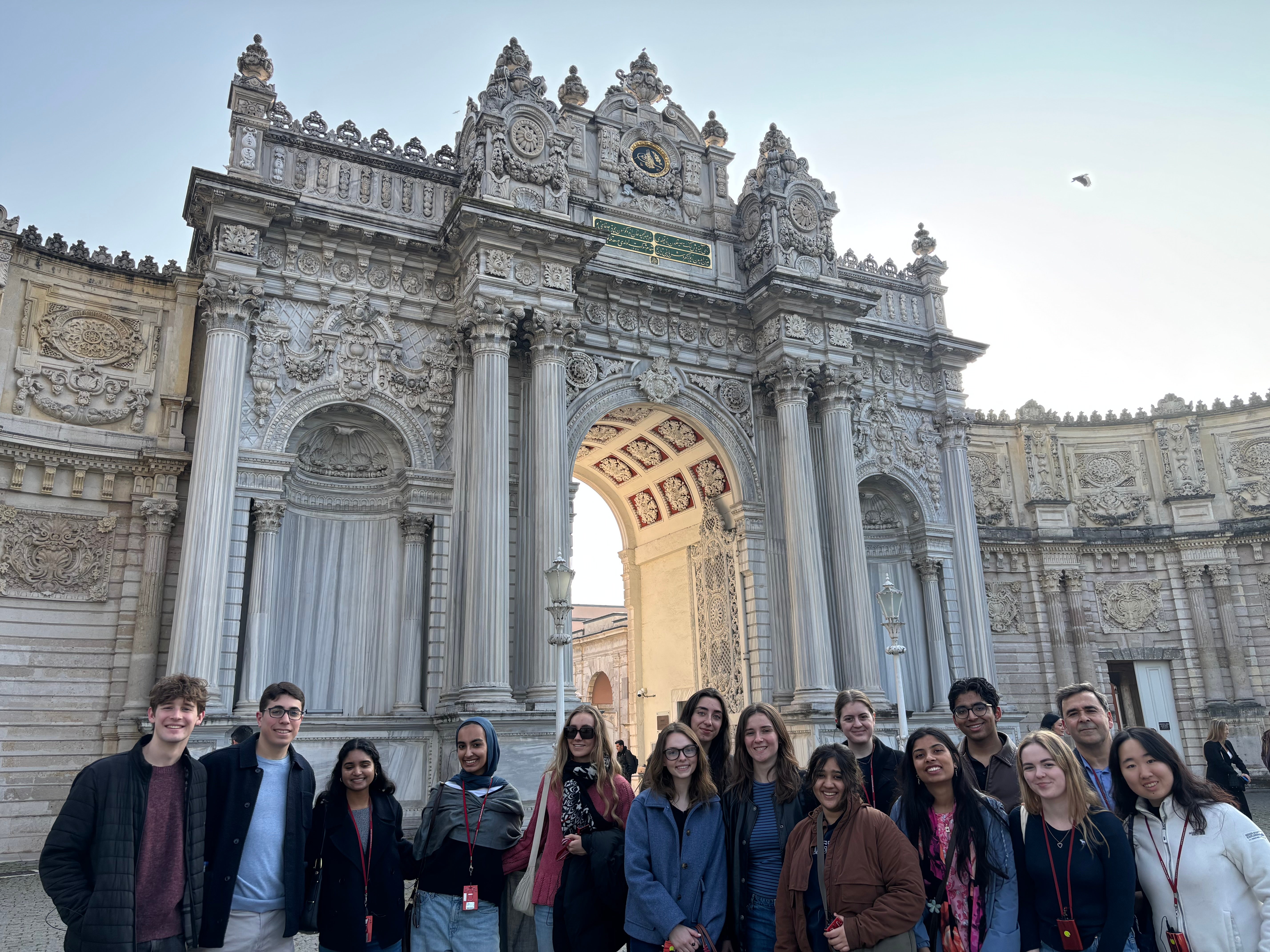 A group of people stands in front of the elaborate, ornate gates of Dolmabahçe Palace in Istanbul. The gates feature detailed carvings and columns. The sky is clear with a hint of blue.
