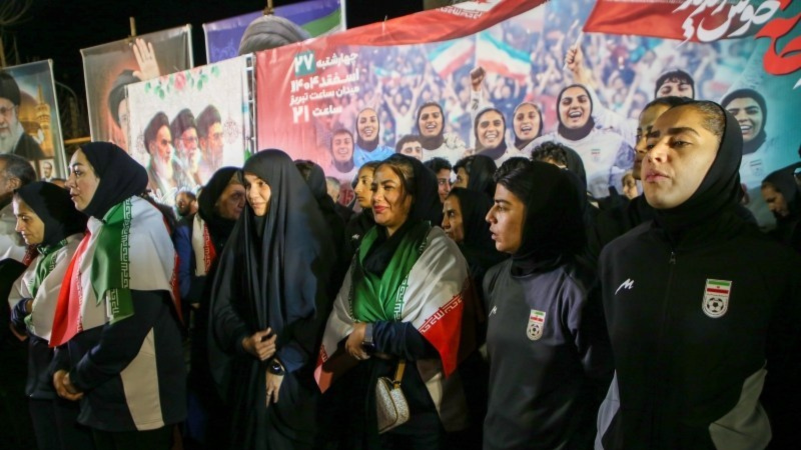 Five Iranian women stand in front of a large billboard of the Iranian women's soccer team.