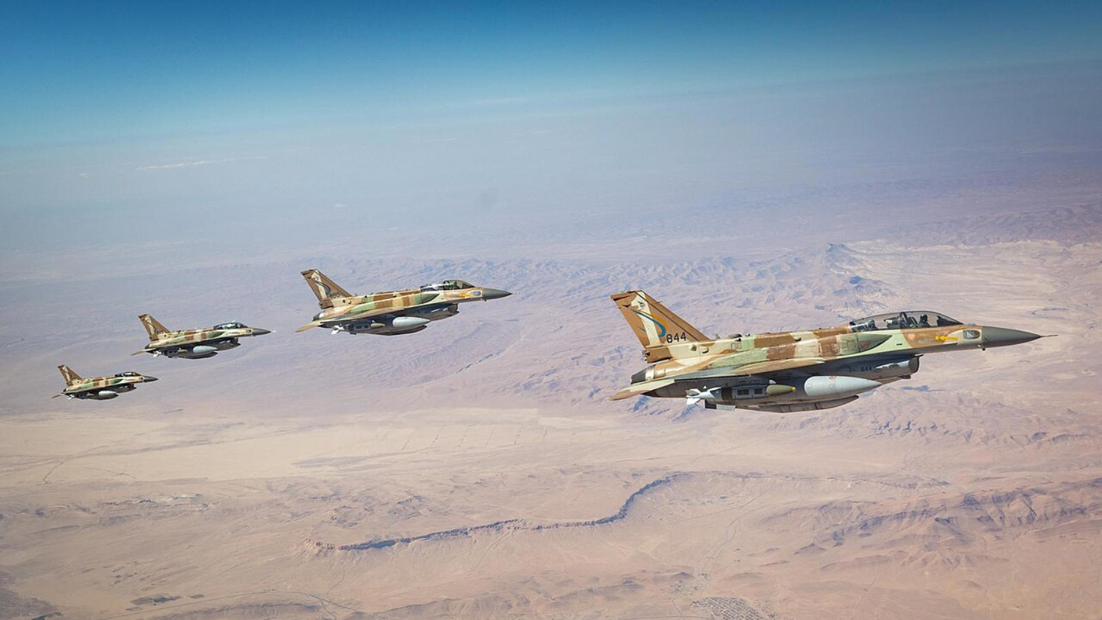 Four Israeli fighter jets with desert camouflage paint fly over a desert landscape.