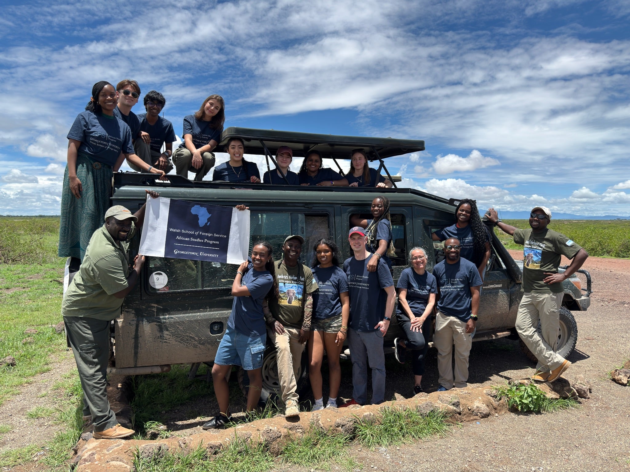 A group of people gathered around a safari vehicle in an open savannah landscape under a cloudy sky. They're holding a banner that reads "African Wildlife Foundation's Maasai Mara Project, Government of Kenya." Some people are standing on the vehicle while others are beside it. The scene conveys a sense of unity and collaboration in a natural setting.