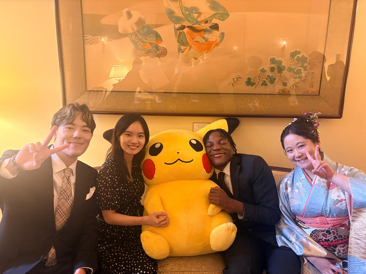 A group of four people are sitting on a couch, smiling and making peace signs. In the center, they hold a large Pikachu plush toy. The setting appears to be indoors, decorated with a framed artwork and soft lighting. One person is wearing a traditional robe with floral patterns.