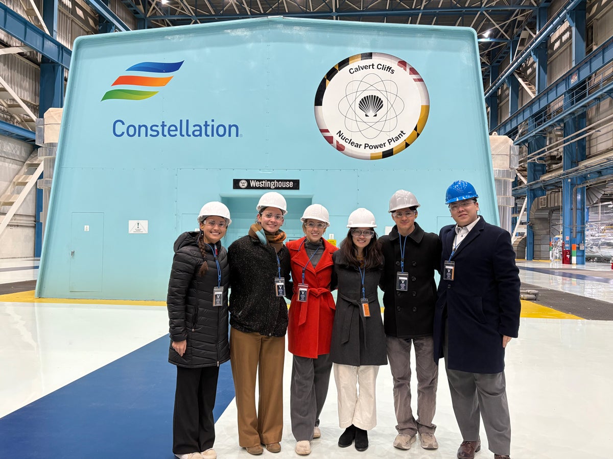 A group of six people wearing safety helmets and badges stand inside a large industrial building. Behind them, there is a sign for the Calvert Cliffs Nuclear Power Plant and the Constellation logo. The floor is a smooth, glossy surface, and the space is brightly lit.