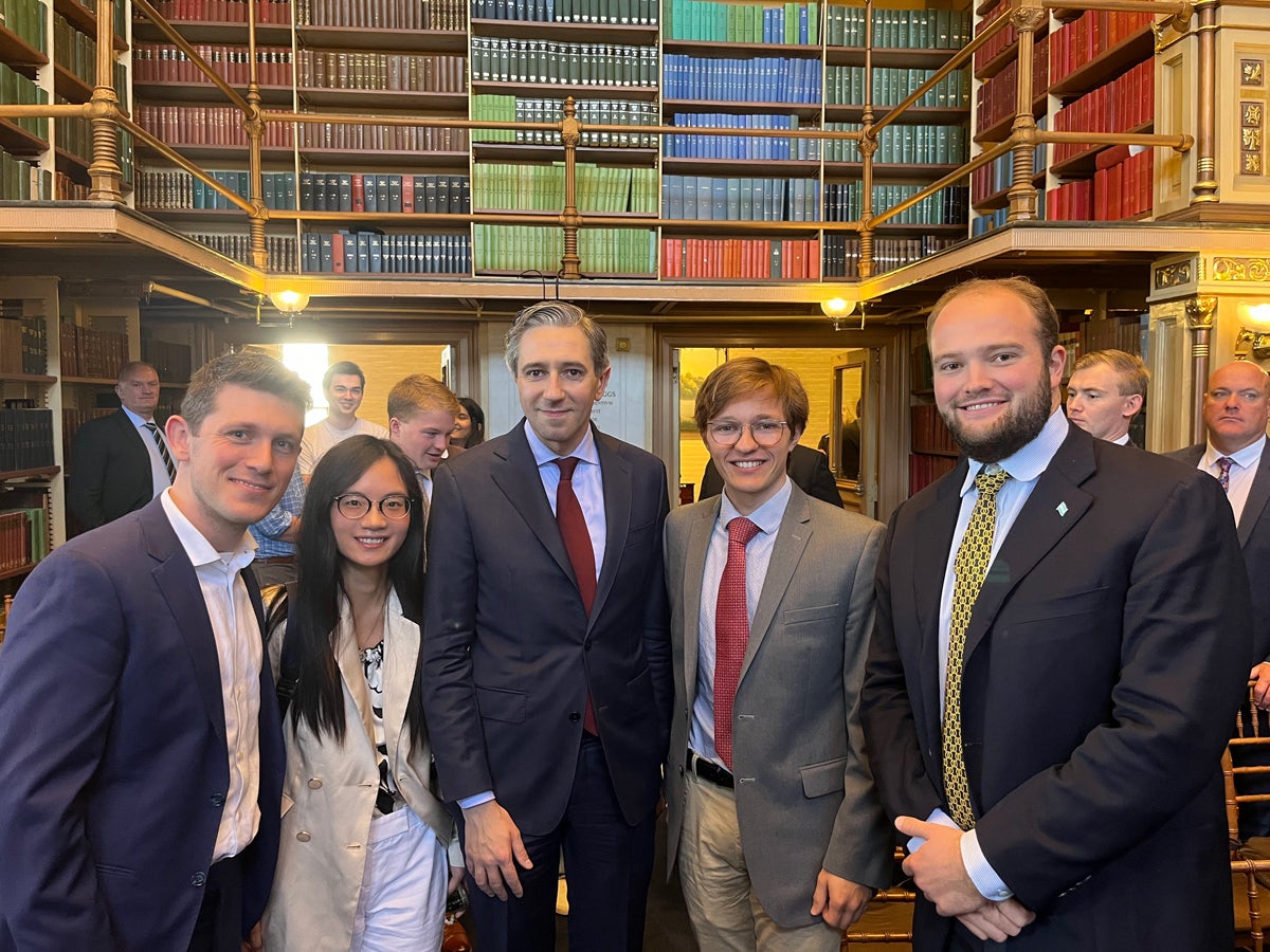 A group of people are standing in an elegant library with tall bookshelves filled with colorful books. Among the group is a person wearing a suit and tie, and others dressed in formal and semi-formal attire. The setting suggests an academic or professional event.