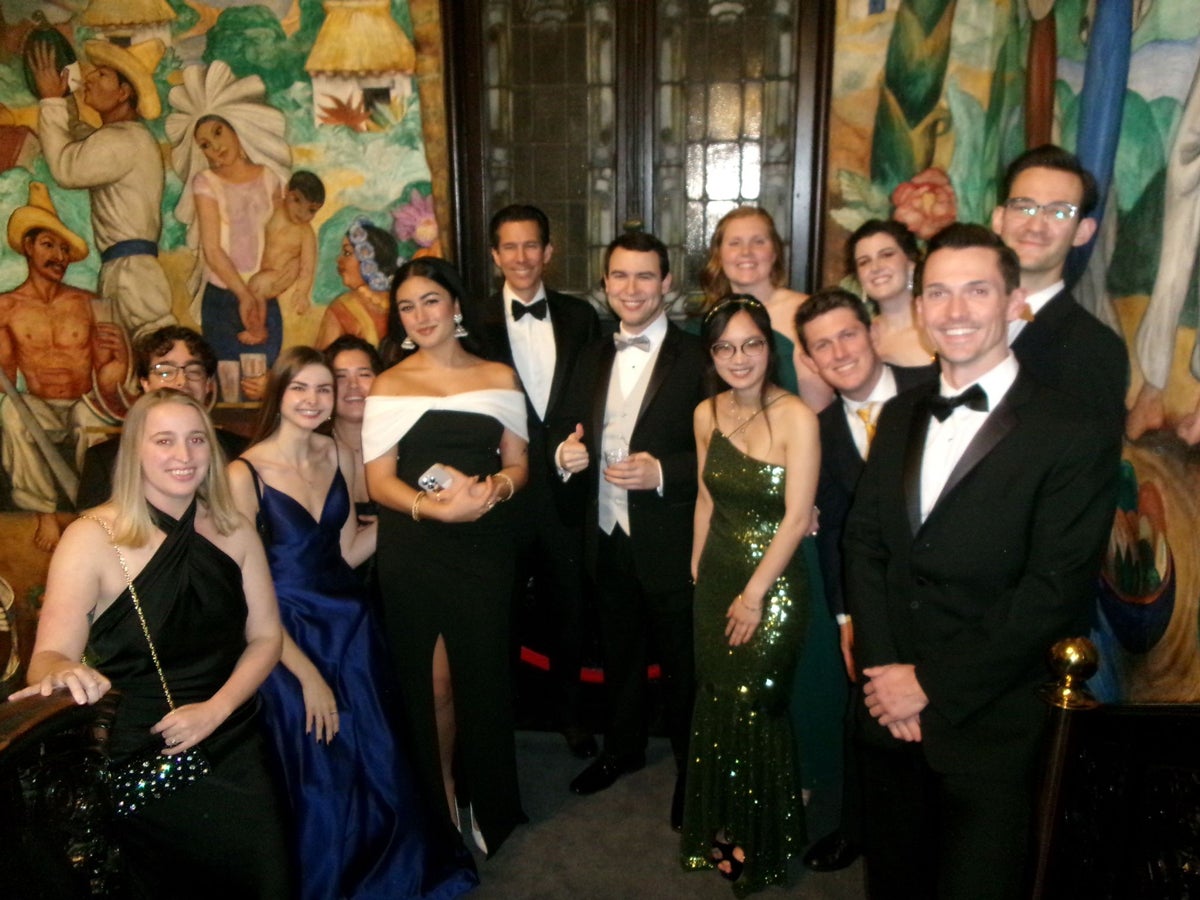 A group of people in formal attire stands in an ornate room with colorful murals by Diego Rivera on the walls. Some hold drinks, and they are smiling at the camera.
